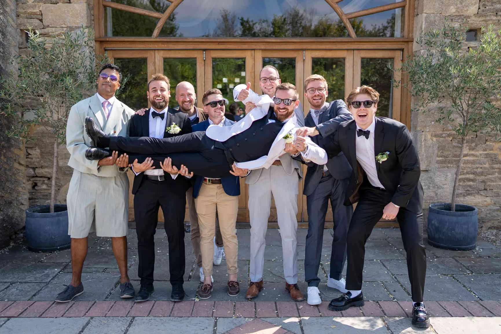 Group of men in suits celebrating at a wedding, lifting Groom in a tuxedo in the air in front of a building with glass doors and stone walls during Oxleaze Barn Wedding