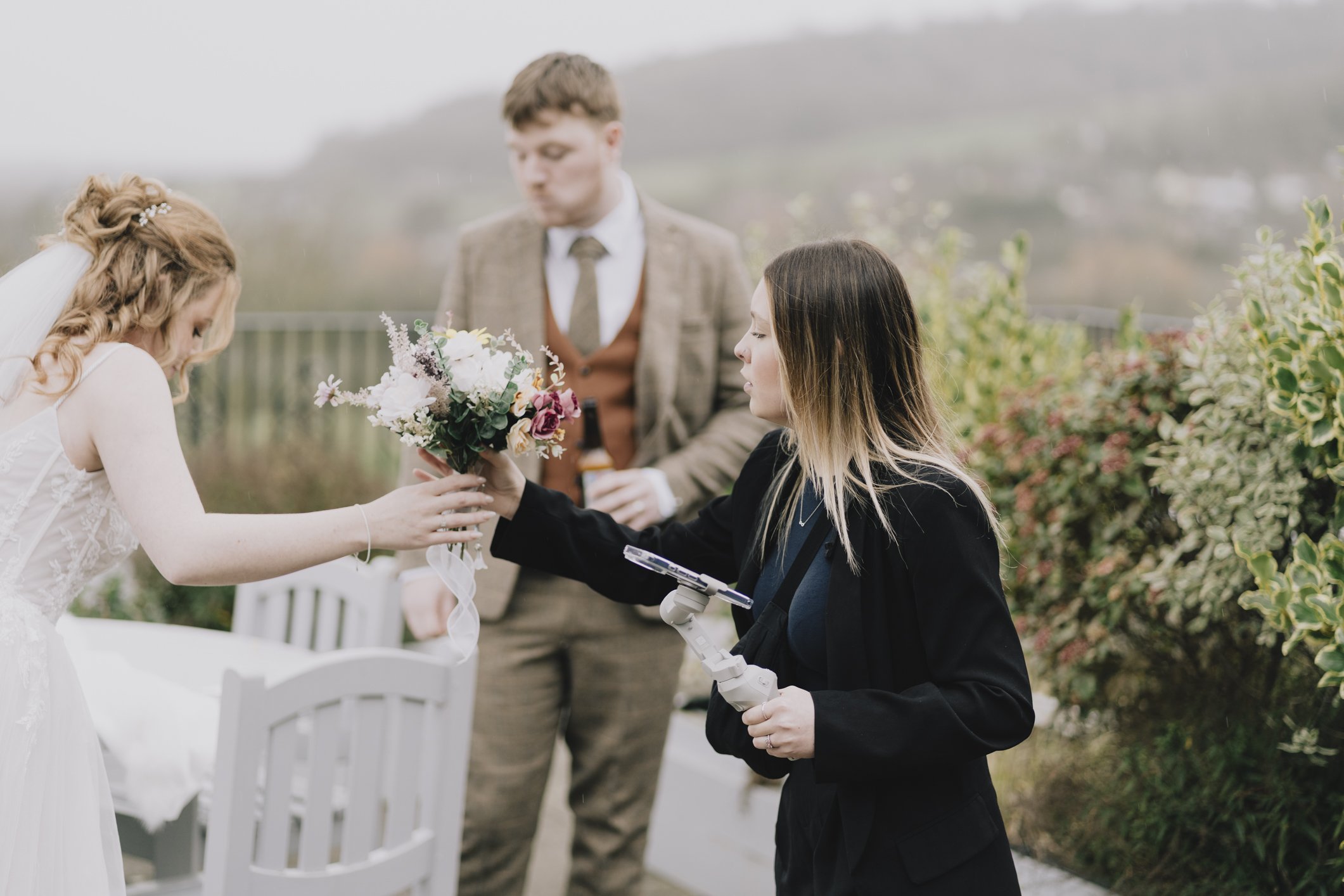 A bride receiving a bouquet of flowers from a The Social Bell Content Creator at Webbington Terrace with a man holding a drink in the background, surrounded by greenery and outdoor scenery.
