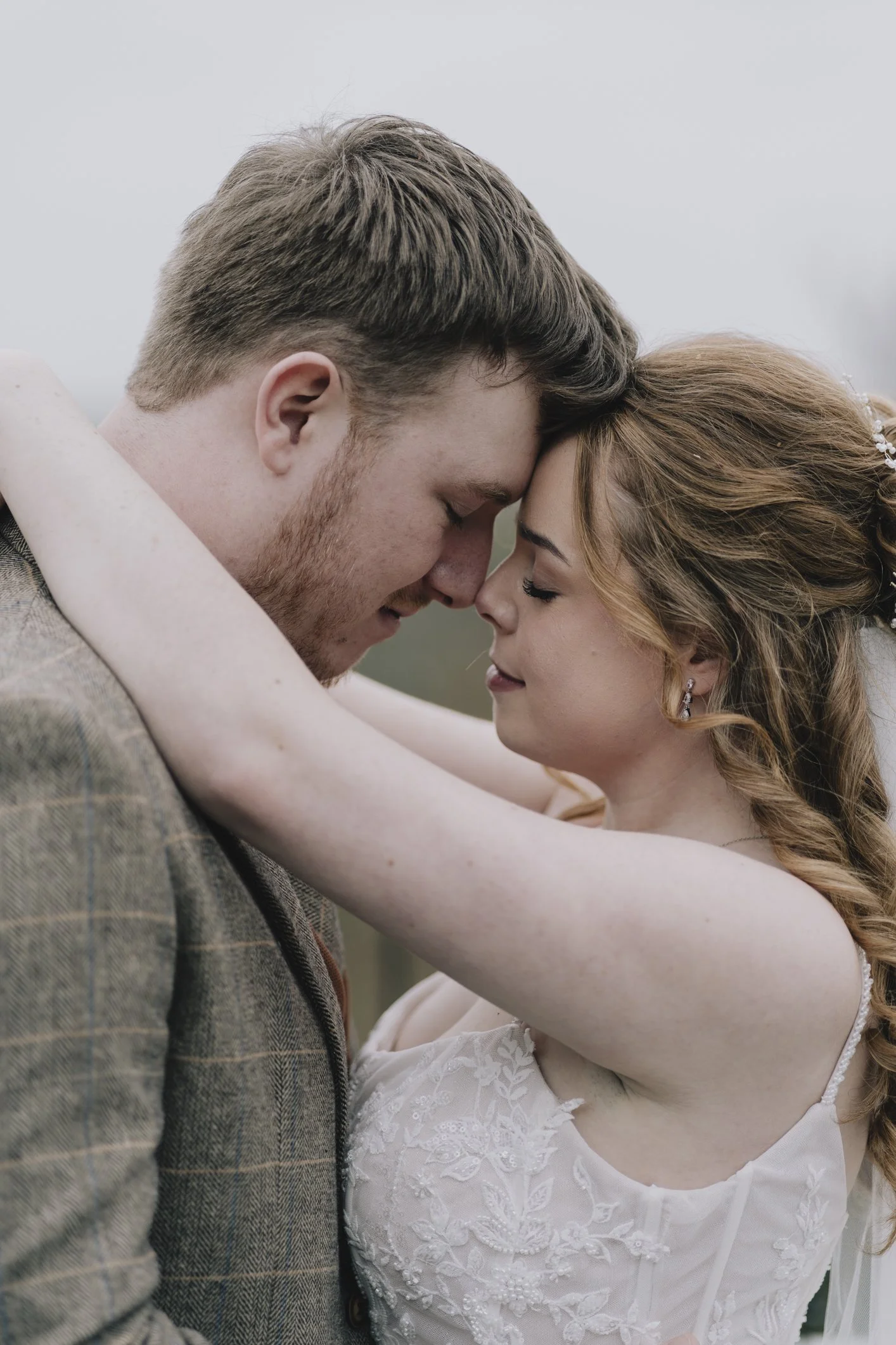 A couple embracing with their foreheads touching, outdoors on a cloudy day, dressed in wedding attire.