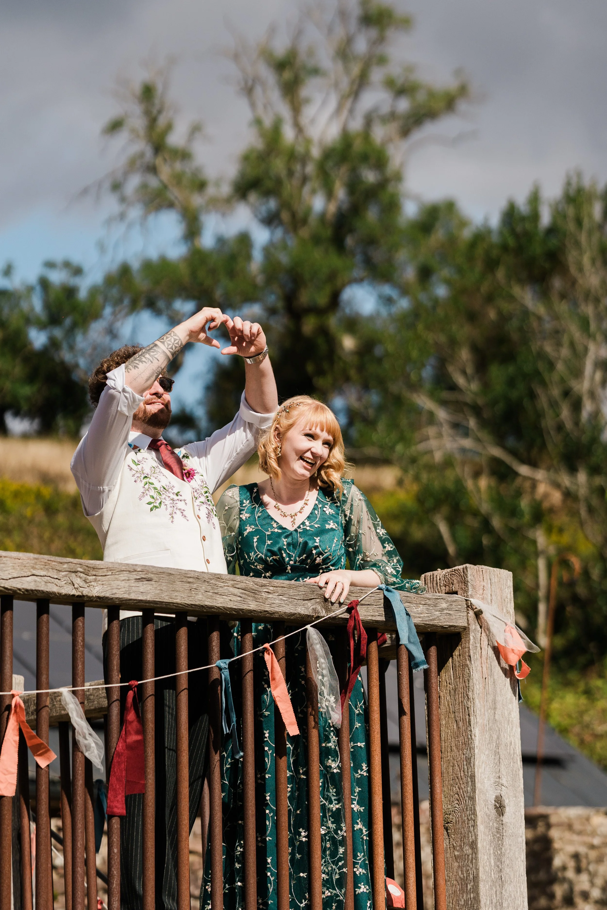 The bride and groom n on a wooden balcony celebrating their wedding both smiling, both dressed in floral clothing, making a heart shape with their hands, decorated with colorful fabric strips.