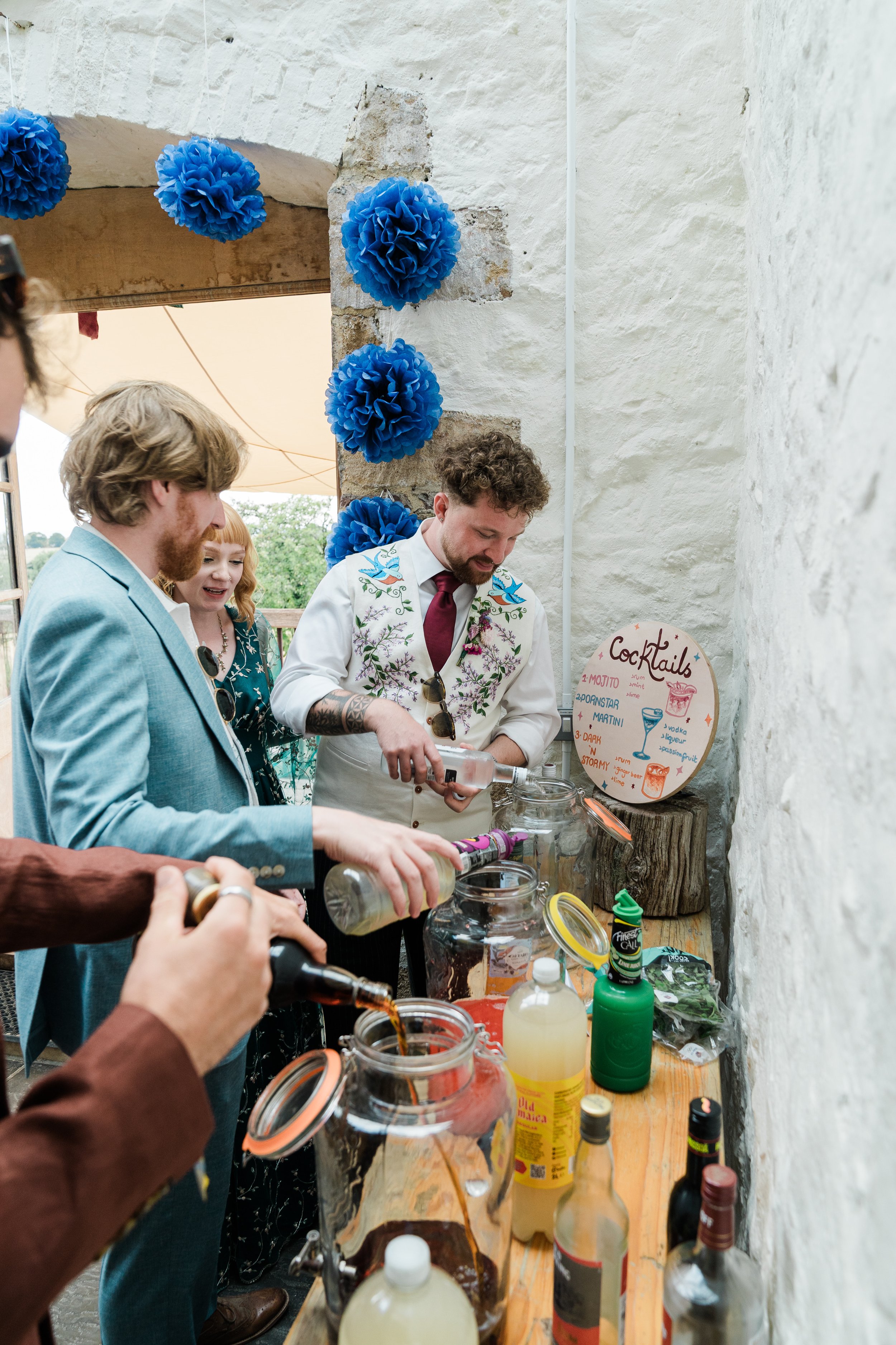People at a bar or drink station with various bottles and beverages, some pouring drinks, in a rustic setting with white walls and blue paper pom-pom decorations.