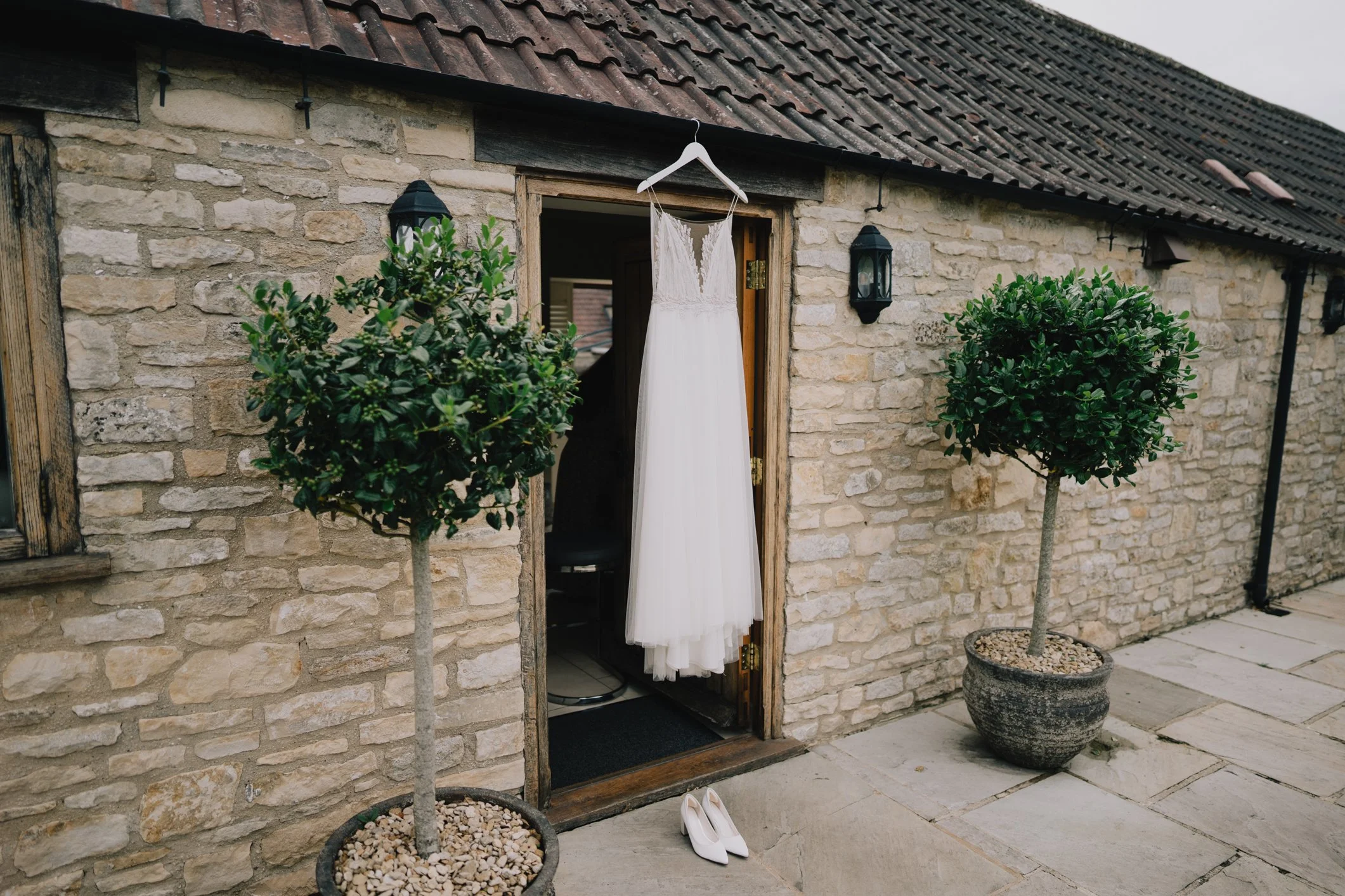 A white wedding dress hanging on a hanger outside a stone building. Two potted trees are on either side of the dress, and a pair of white high-heeled shoes are on the ground below it near Tetbury