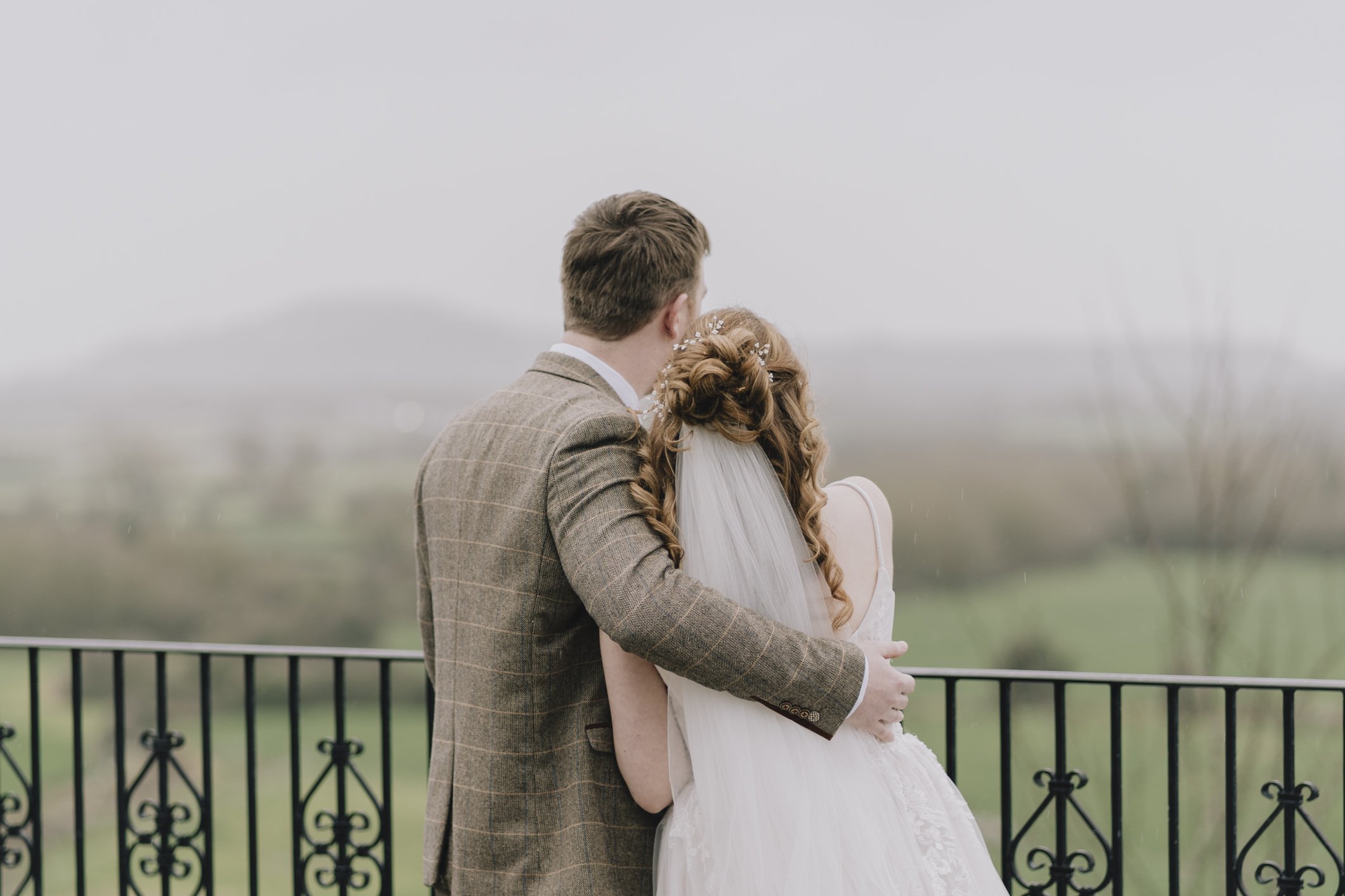 A newlywed couple stands on a balcony overlooking a rural landscape with hills in the background on a cloudy day at The Webbington Hotel.