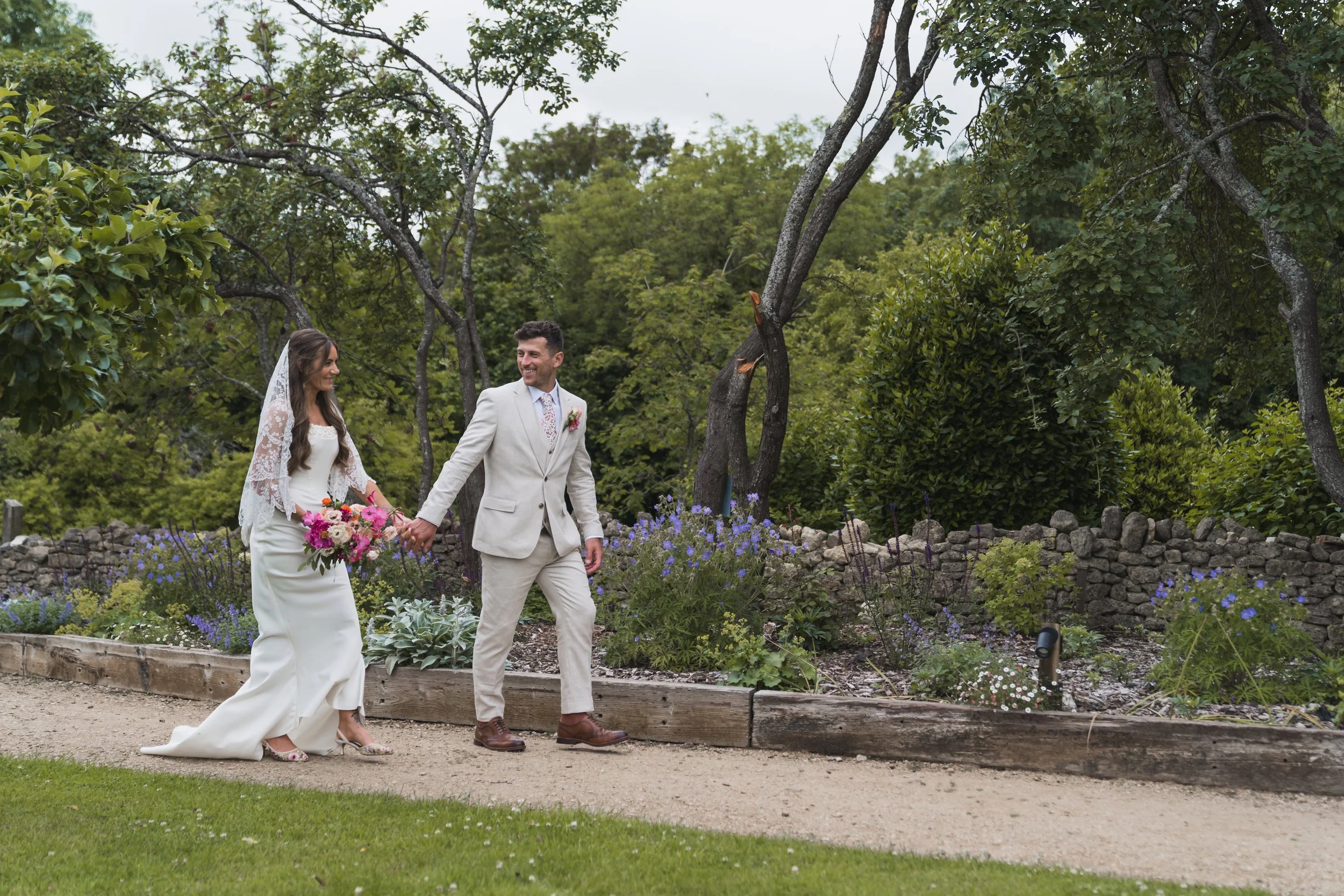 A bride and groom holding hands walking through a garden with purple flowers, trees, and a stone wall in background.