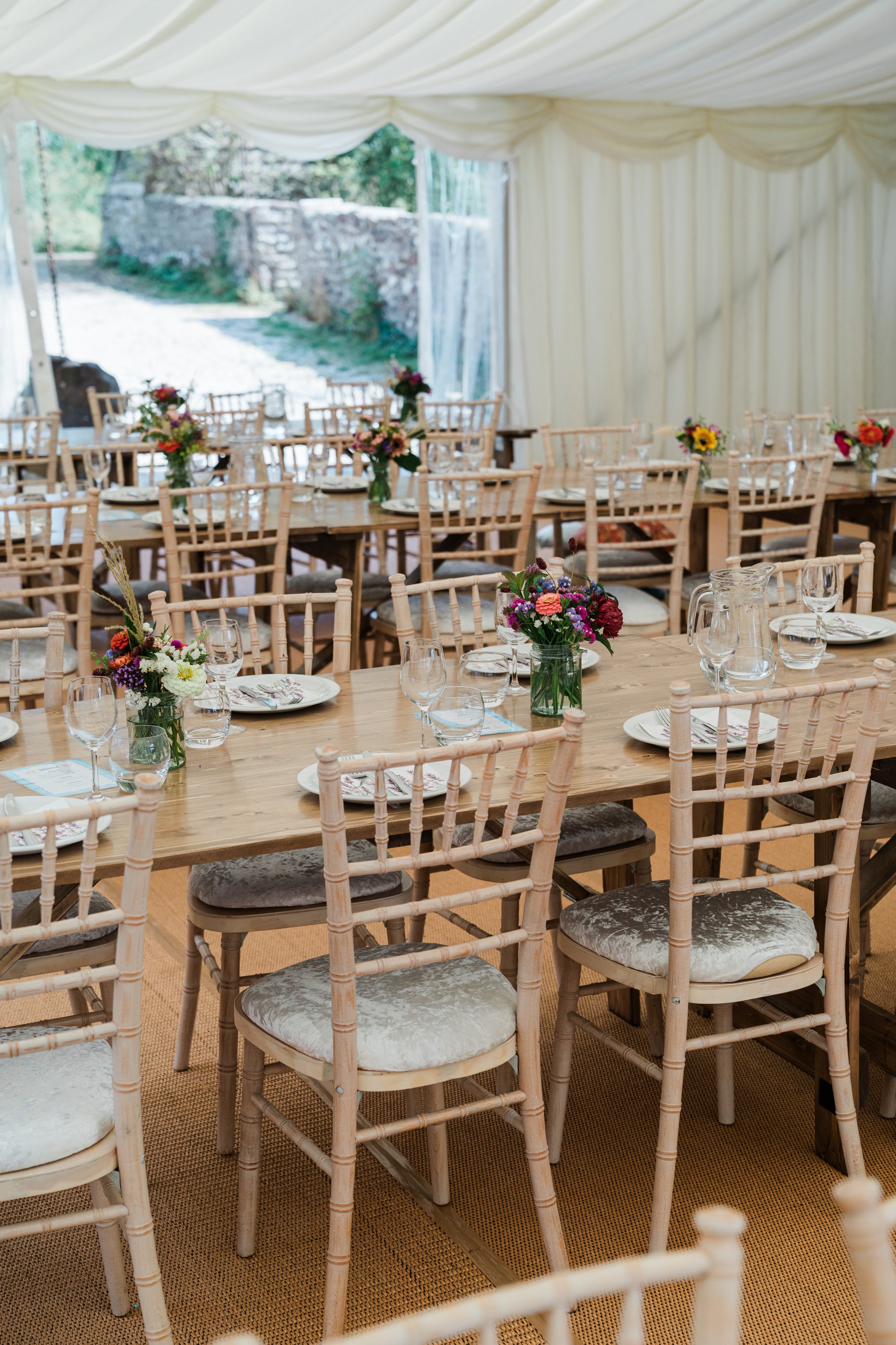 Wedding reception setup with long wooden tables, beige chairs with velvet cushions, and floral centerpieces under a white tent with open sides.