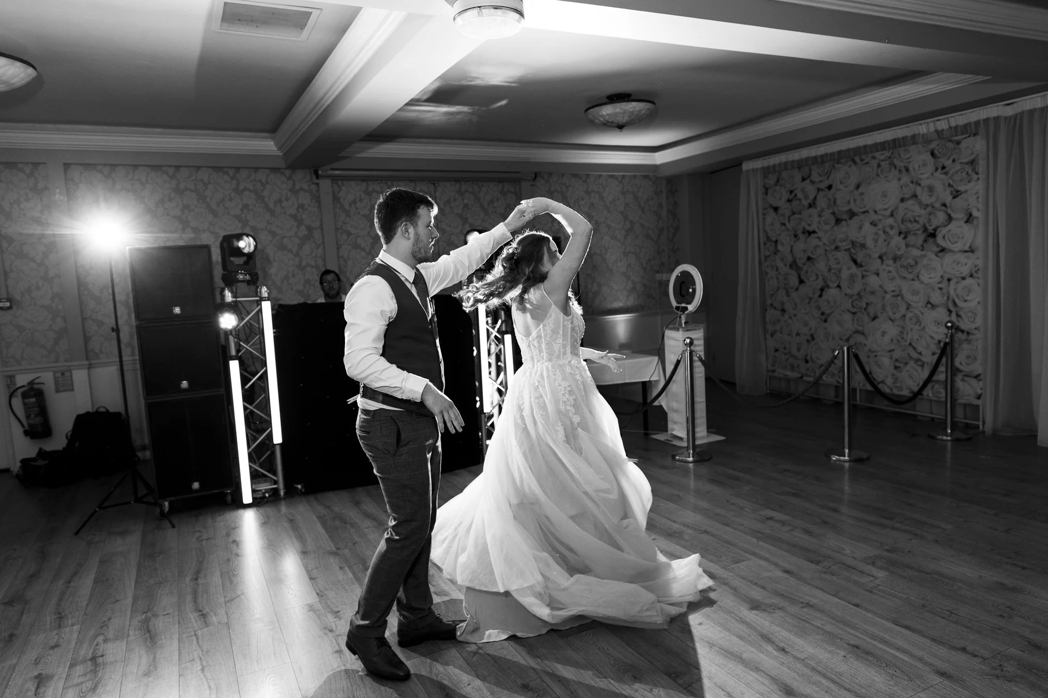 A black and white photo of a bride and groom dancing at their wedding reception. The groom is spinning the bride, who is wearing a flowing wedding dress, in a decorated hall with wooden floors and floral wallpaper.