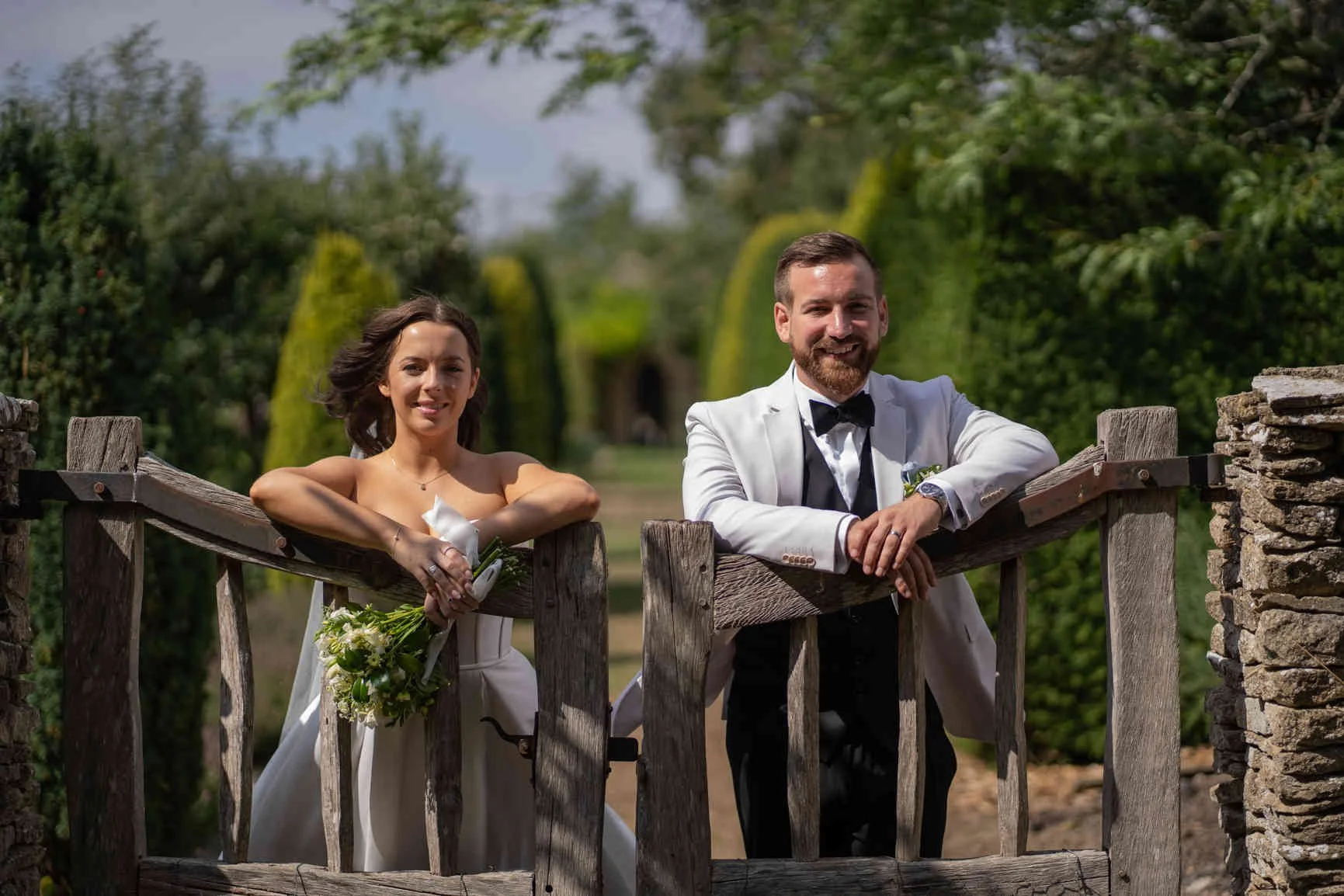 Portrait of a newlywed couple leaning on a rustic wooden gate outdoors, surrounded by greenery and trees, smiling at the Gloucester Wedding Photographer.