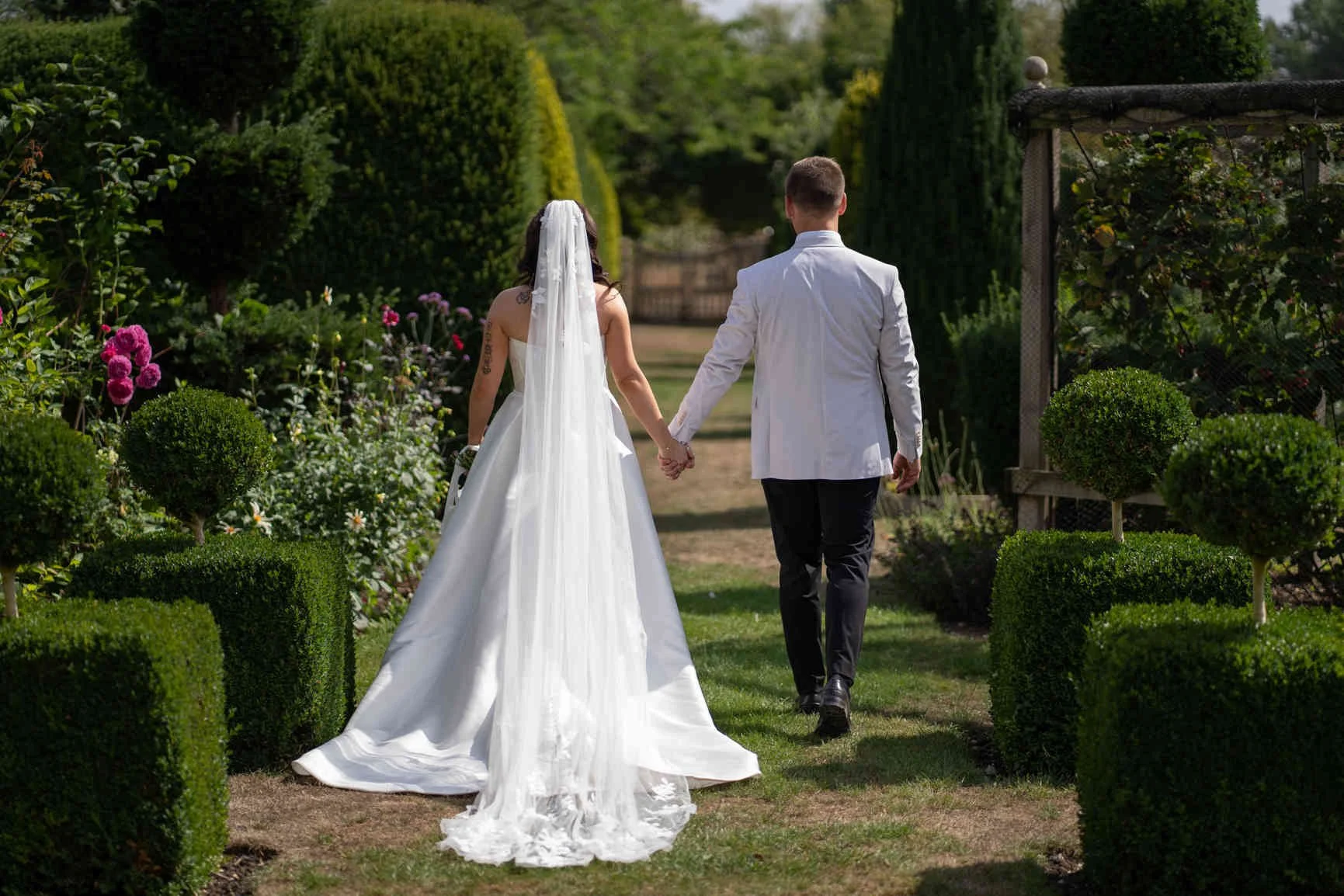 A bride and groom walk hand in hand through a Oxleaze Barn pathway with manicured bushes and colorful flowers on either side, holding hands and dressed in wedding attire.