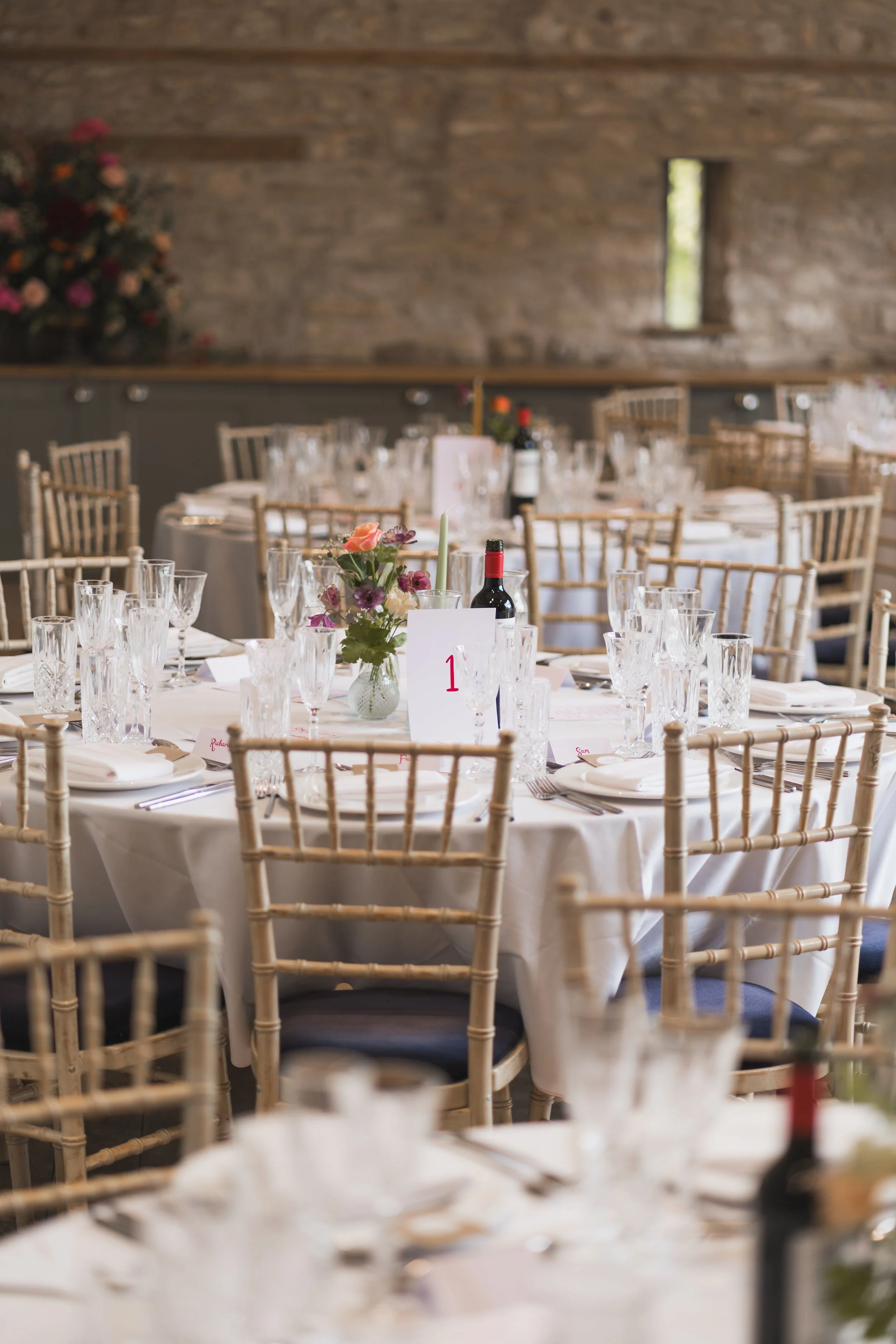 A wedding reception setup with multiple round tables adorned with white tablecloths, floral centerpieces, wine bottles, and glassware, inside a rustic barn with wooden walls and narrow windows.