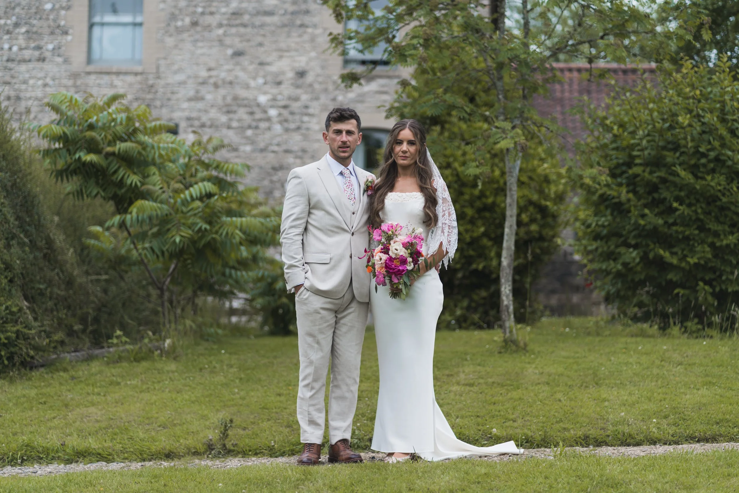 A newlywed couple standing outdoors on a grassy area with some trees and a stone building in the background. The groom is wearing a light-colored suit with a floral tie, and the bride is in a white dress with lace sleeves, holding a bouquet of pink, purple, and white flowers.