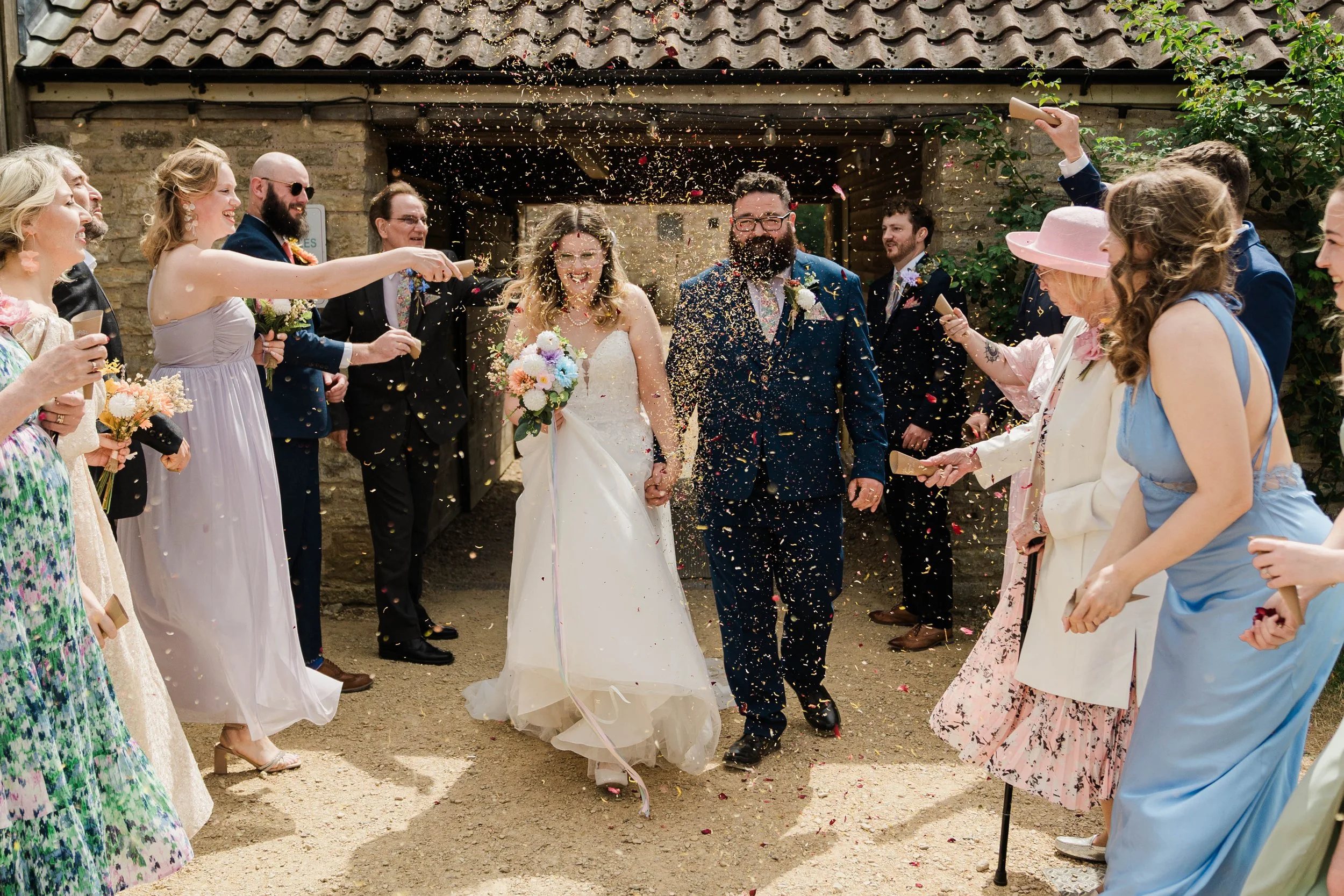 A wedding celebration with guests outside, throwing confetti at a bride and groom holding hands as they smile and walk through a crowd at Folly Farm