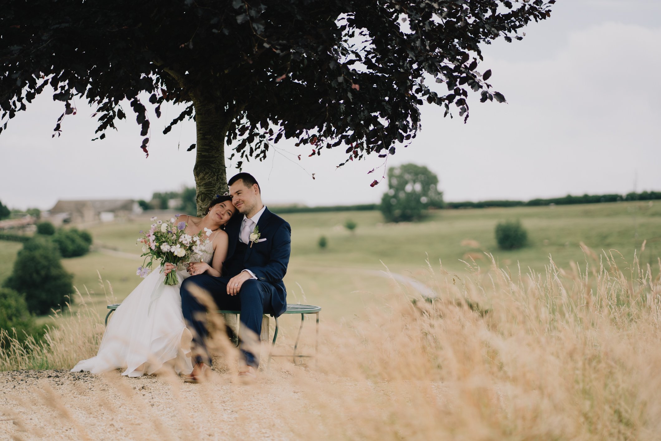 A bride and groom sitting under a tree in a grassy field, with the bride holding a bouquet of flowers, during their wedding photoshoot near Tetbury taking by local photographer