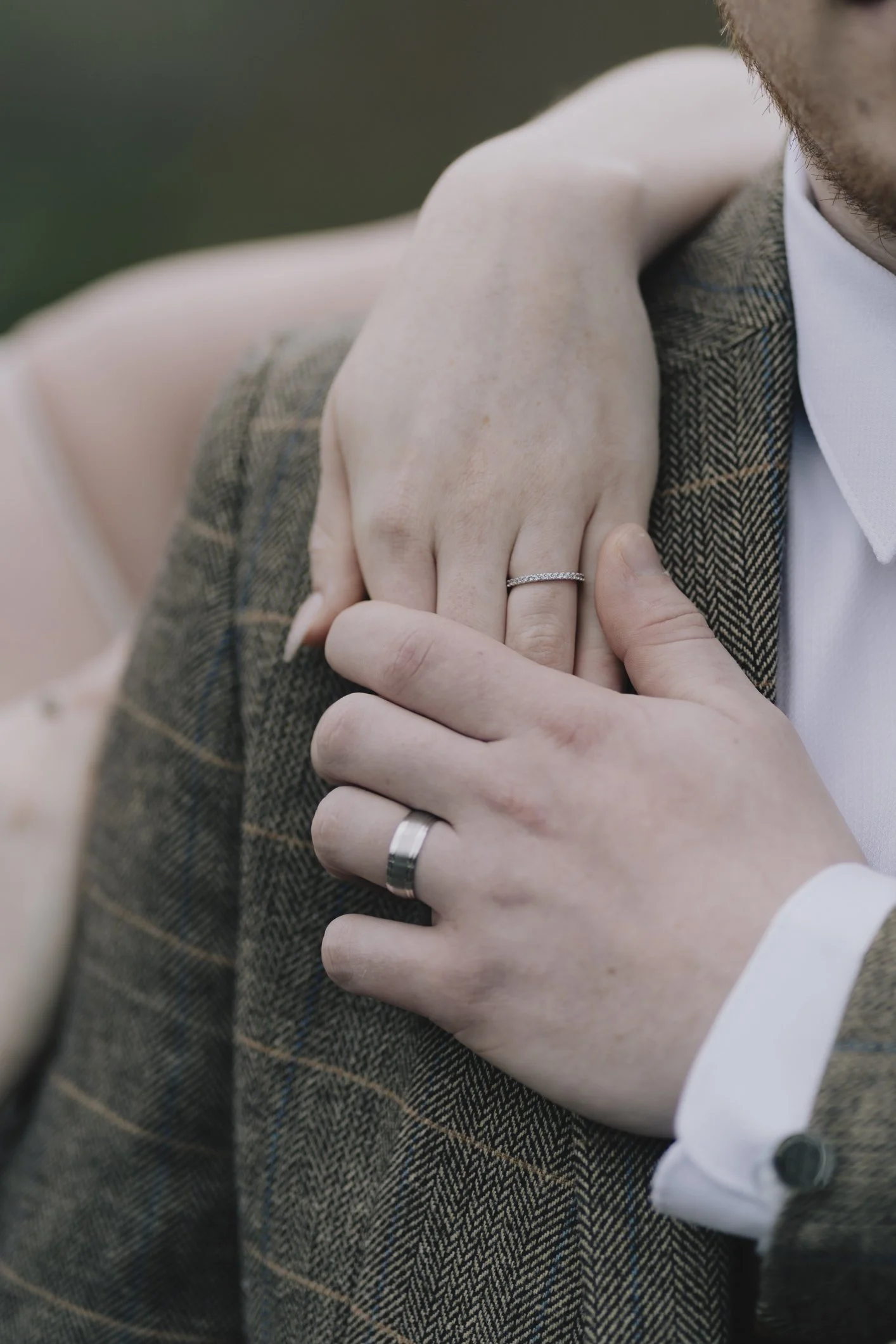 Close-up of a couple's hands, one resting on the other's shoulder, both wearing wedding rings, with formal attire.
