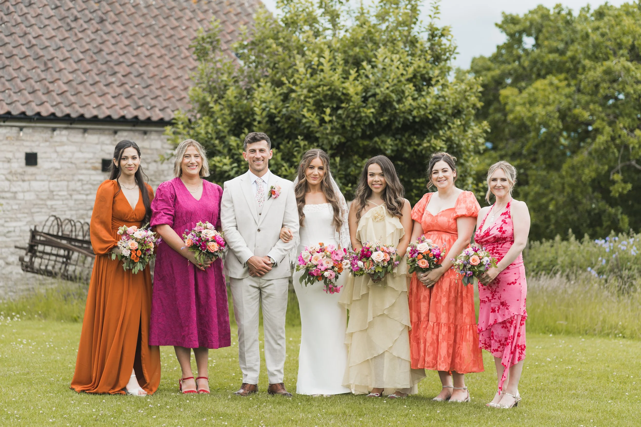A group of women and a man in a wedding dress, standing outdoors on grass with trees and a stone building with a tiled roof in the background. The women are holding bouquets of flowers and are dressed in colorful dresses, while the man is dressed in a light-colored suit.