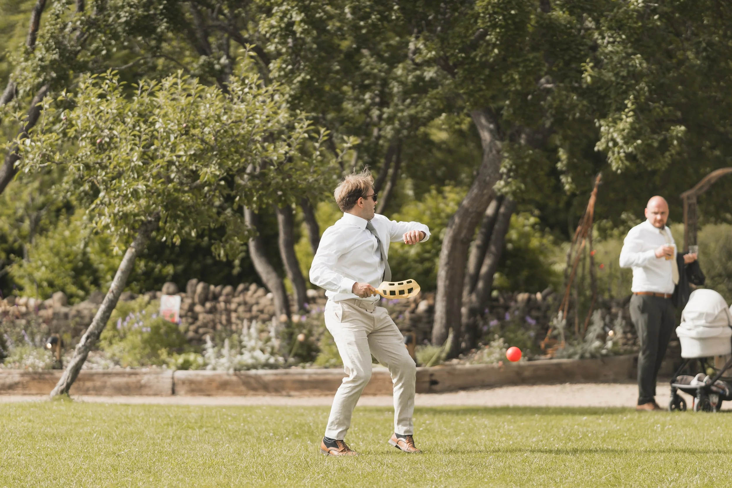 A man in white pants and a white shirt playing croquet outdoors on a grassy area with trees in the background, with another man in a white shirt holding a drink in the background.