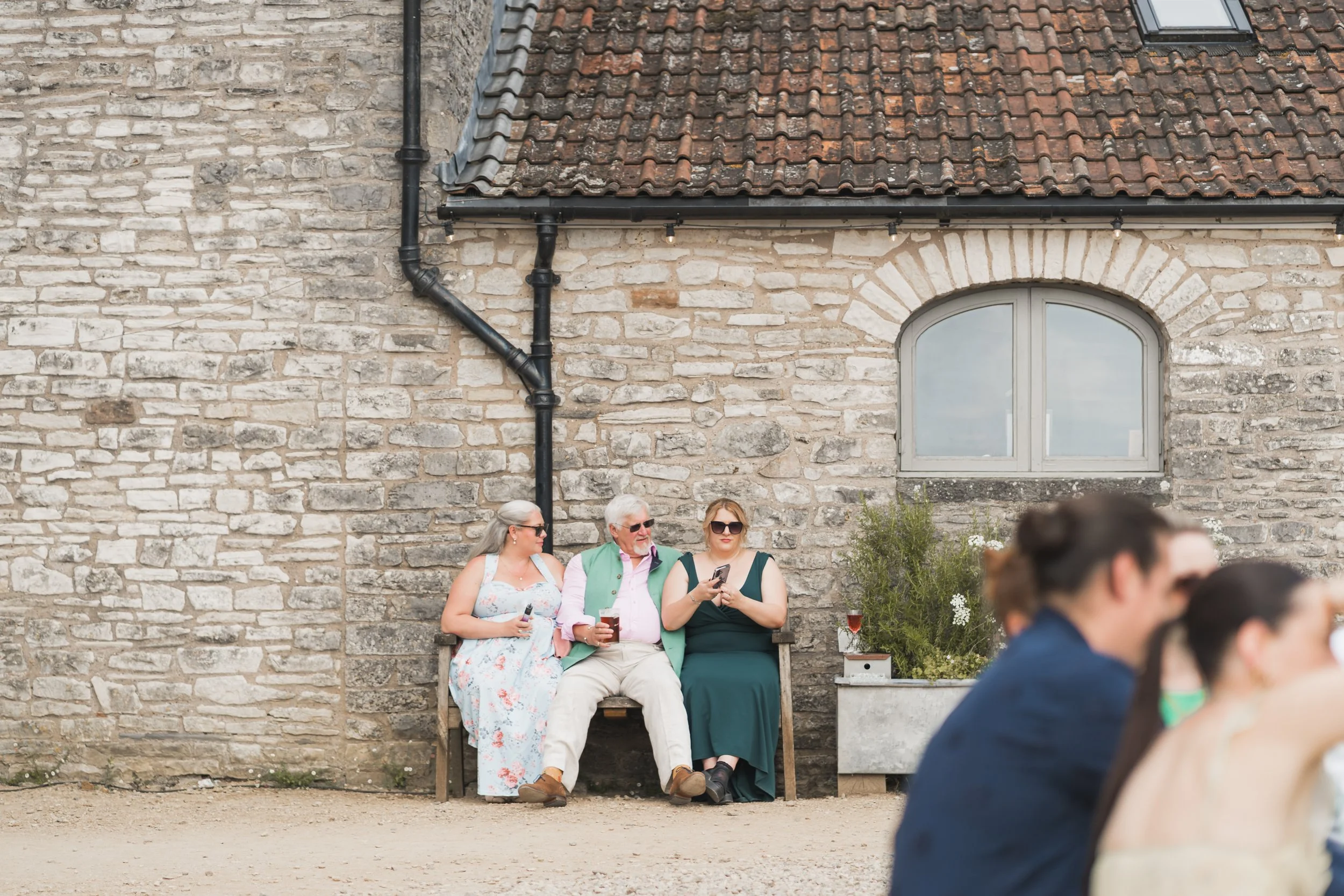 A group of four people sitting on a bench in front of a stone wall, with a man wearing sunglasses and a woman in a dark green dress using her phone, and a woman in a white floral dress and a man in a light green vest engaging in conversation.