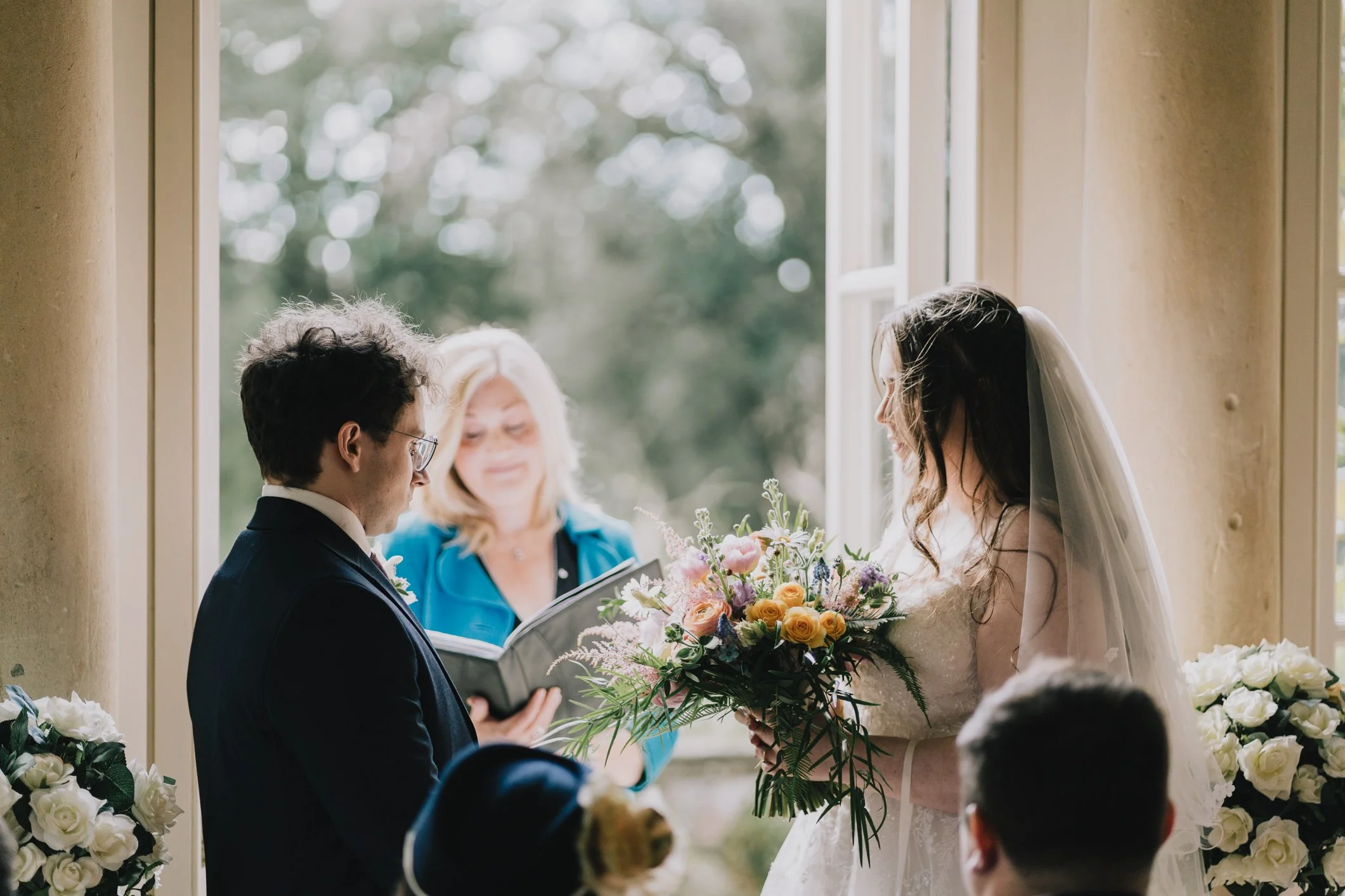 A couple exchanging wedding vows inside Temple of Minerva, Bath , with an officiant holding a book behind them. The bride is wearing a white dress, a veil, and holding a bouquet of flowers.