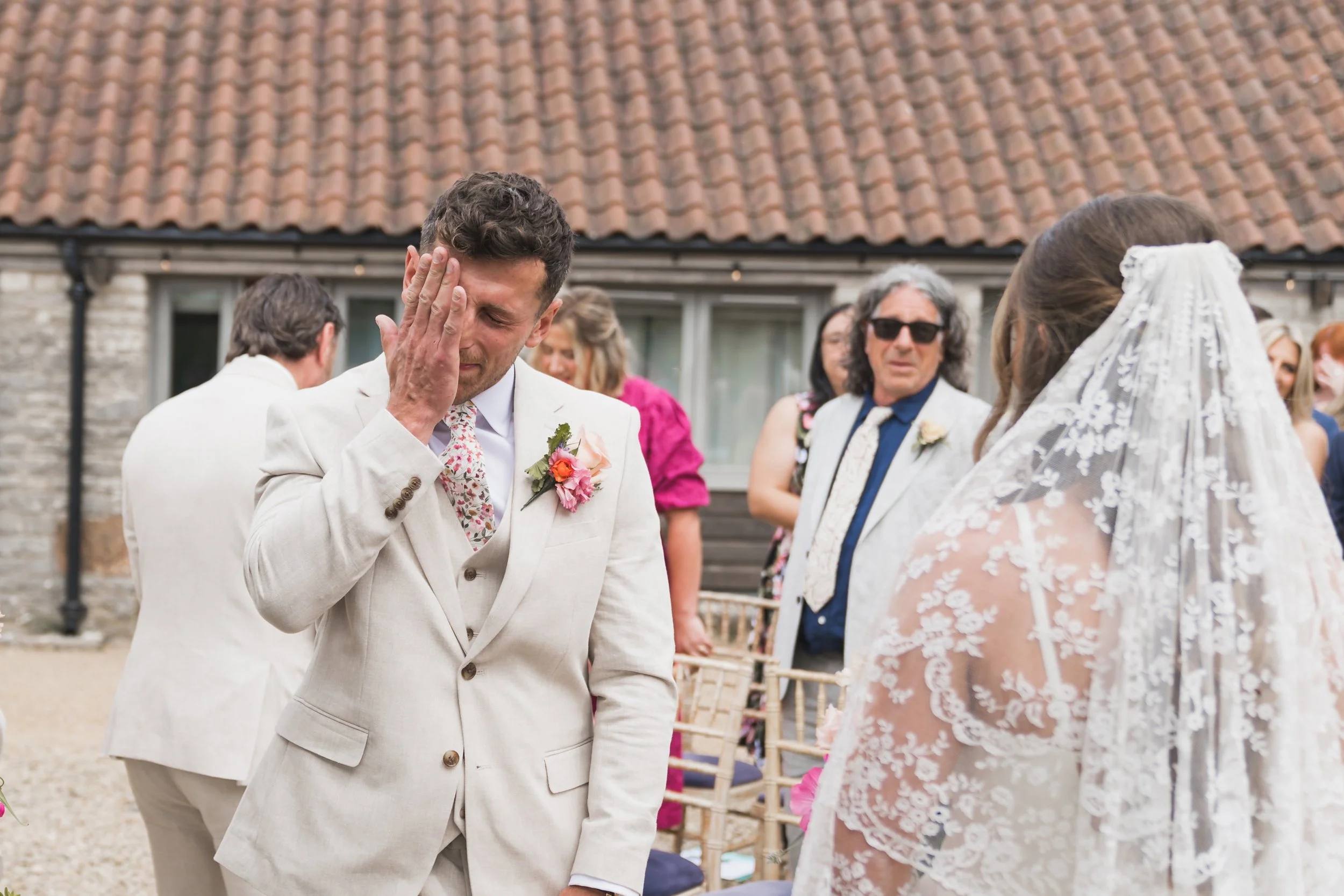 A groom, dressed in a light-colored suit with a floral tie and flower boutonniere, is emotional during a wedding ceremony, touching his face with his hand, while a bride in a lace wedding dress faces him, with guests in the background.