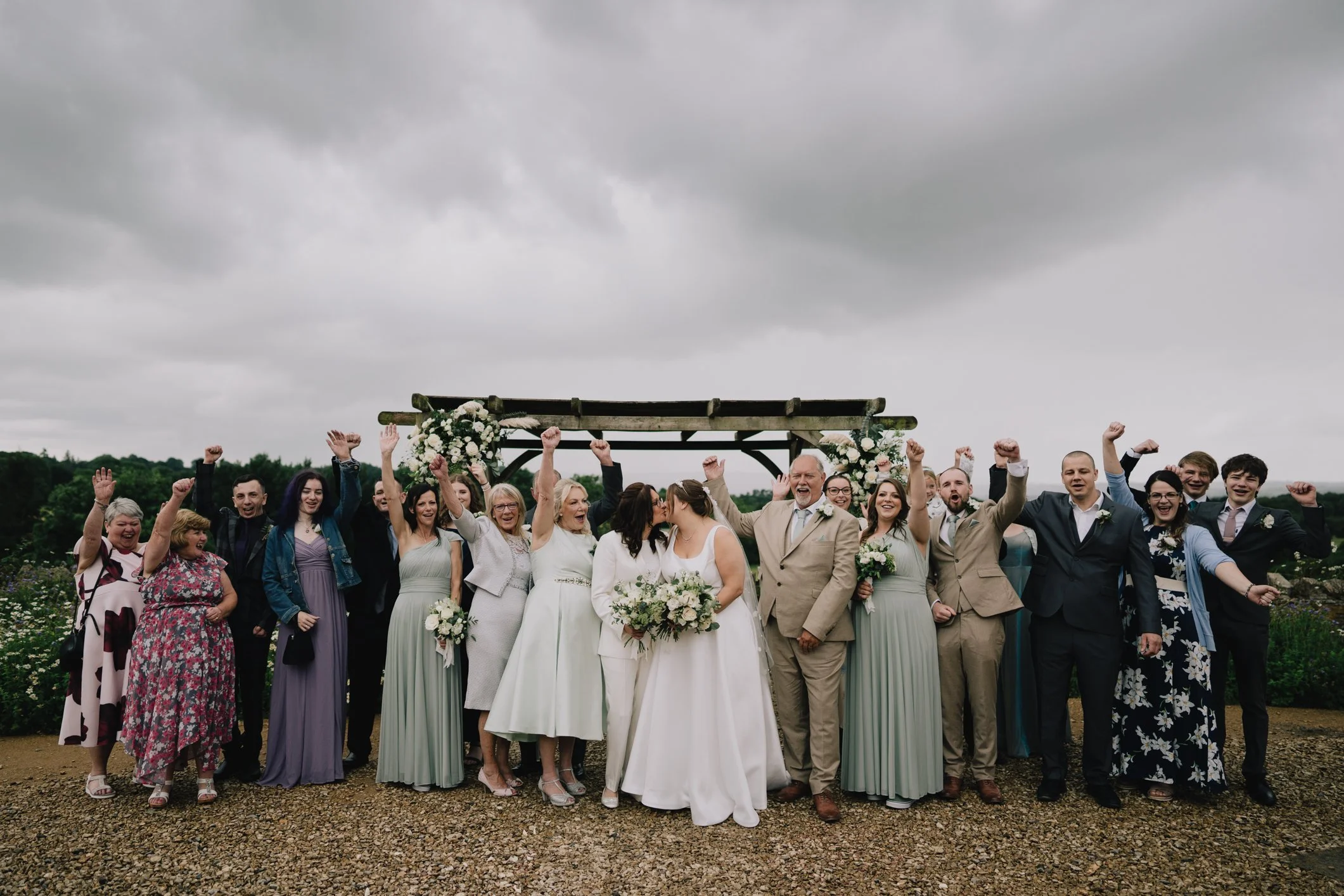A group of people at a wedding celebration outdoors under a cloudy sky, with some raising their fists and others smiling and looking at each other.