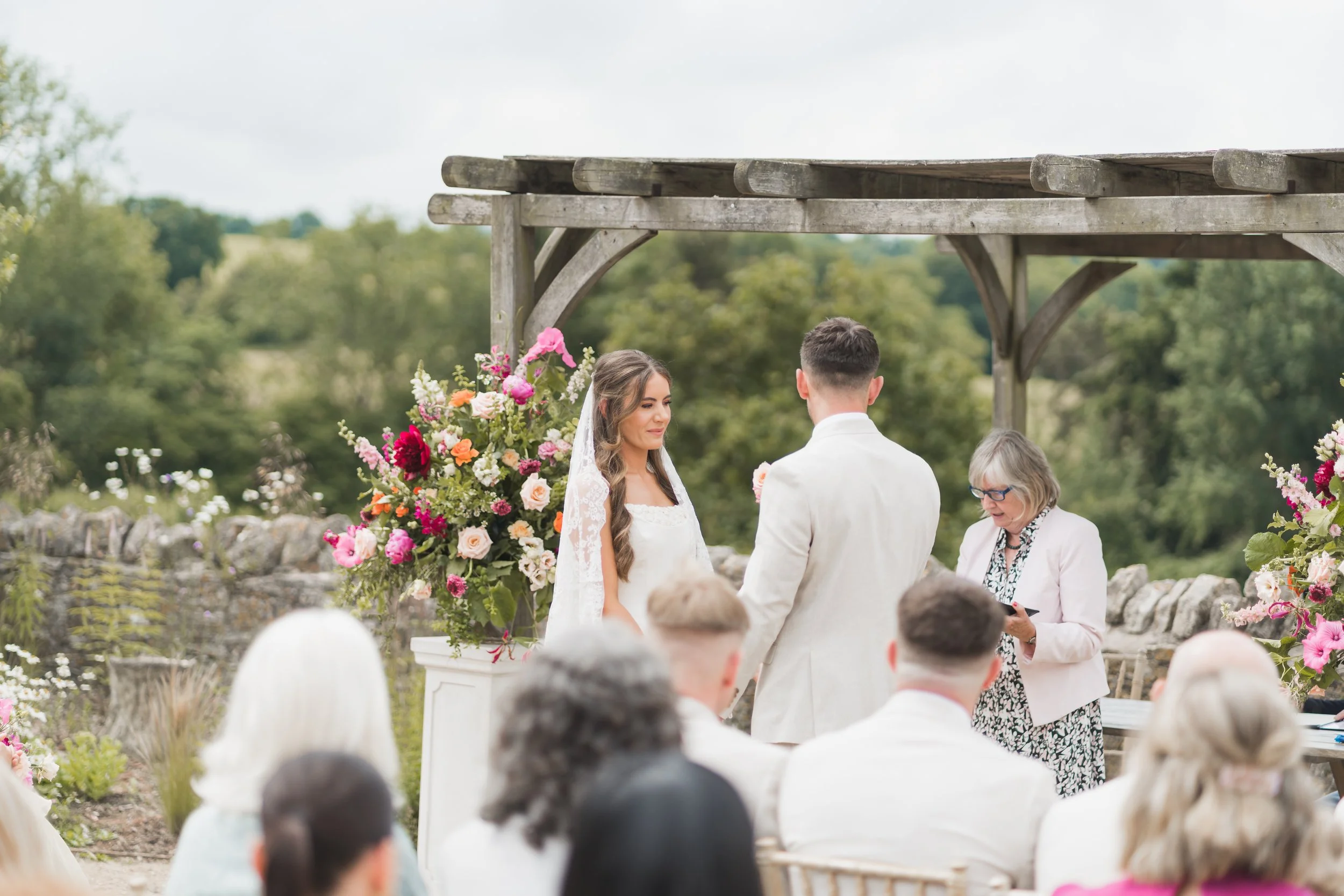 A couple is getting married outdoors at Folly Farm near Bristol, standing before a wedding officiant under a wooden structure with flowers on it, during daytime with green trees in the background.