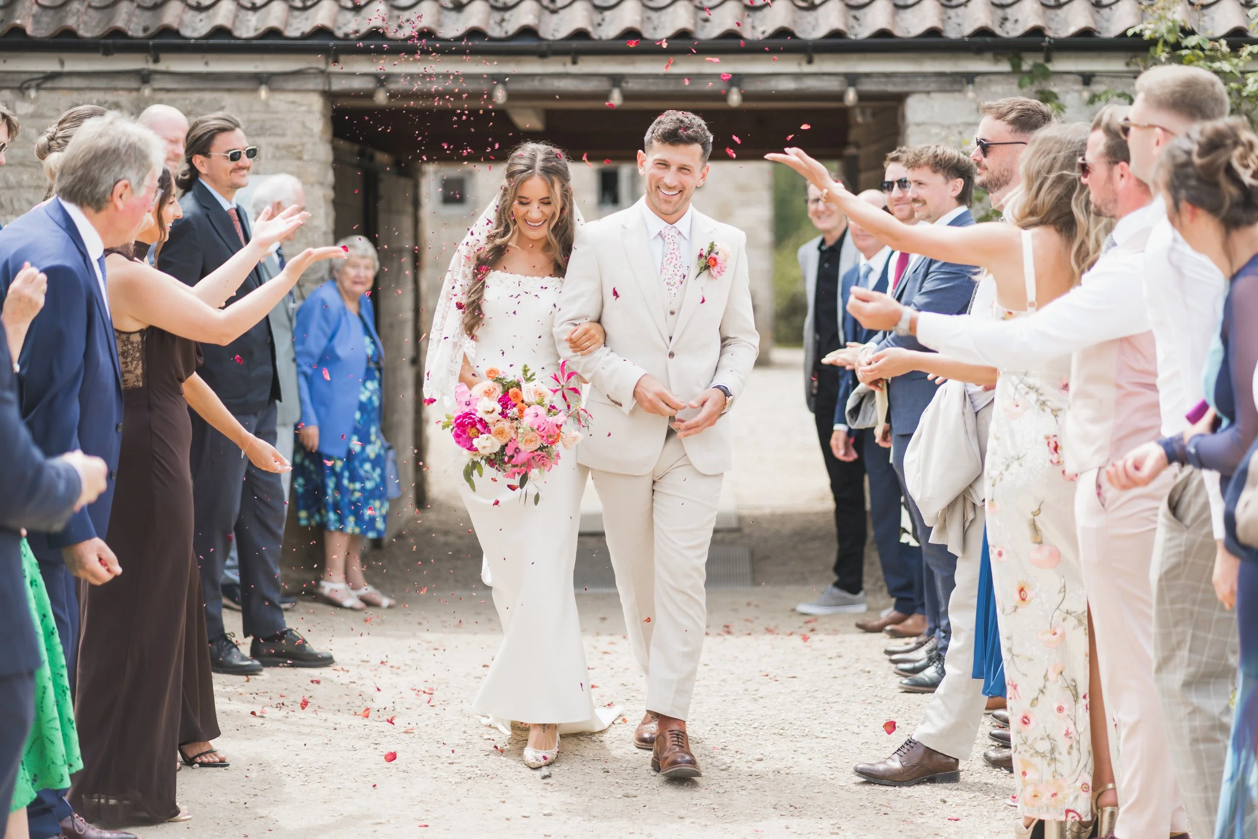 A newlywed couple walks through a path lined with guests throwing flower petals during their wedding celebration outside a stone building.