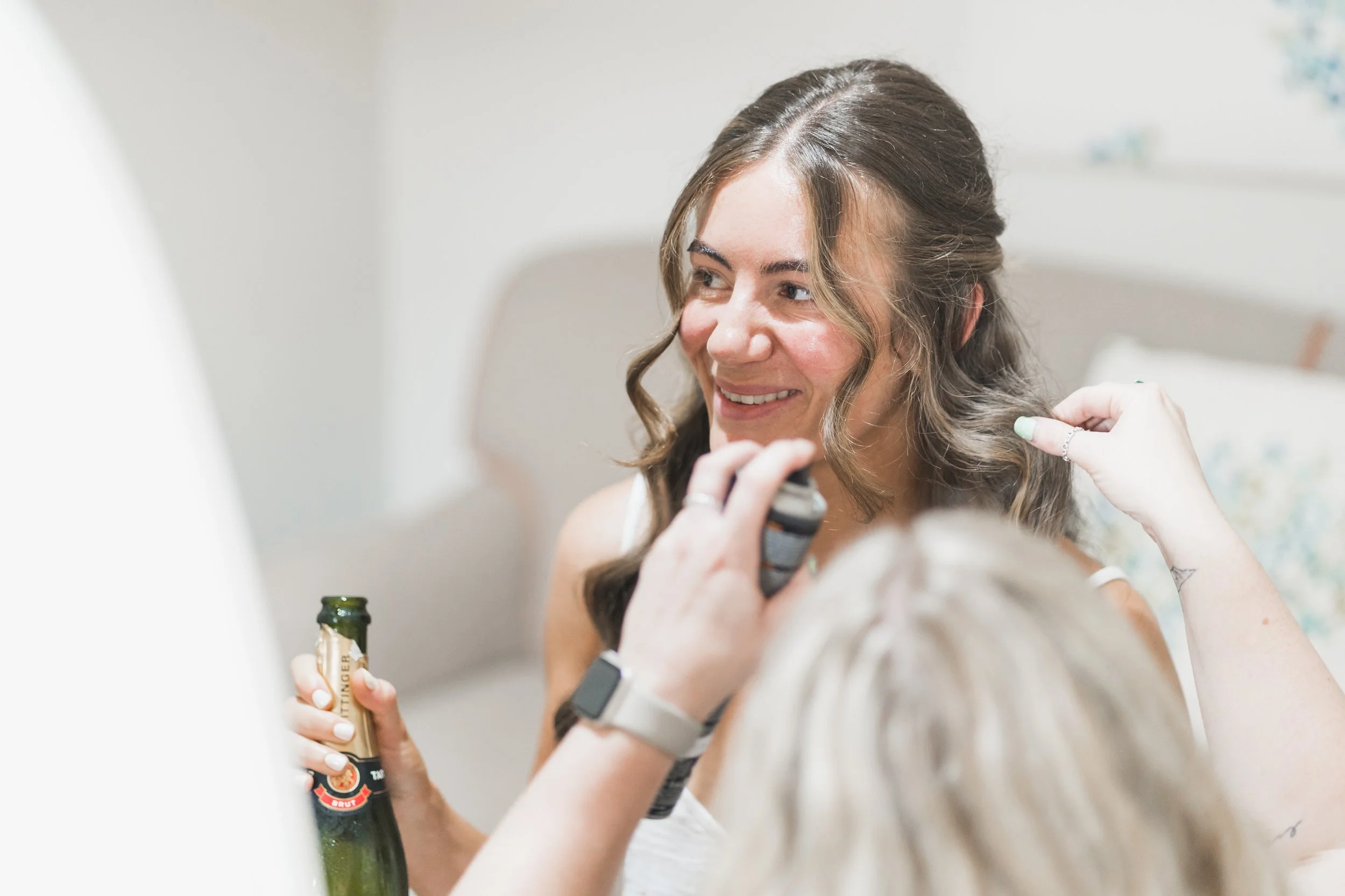 A woman with long wavy hair smiling as someone gives her a haircut in a bright room. She is holding a bottle and wearing a ring and a bracelet.
