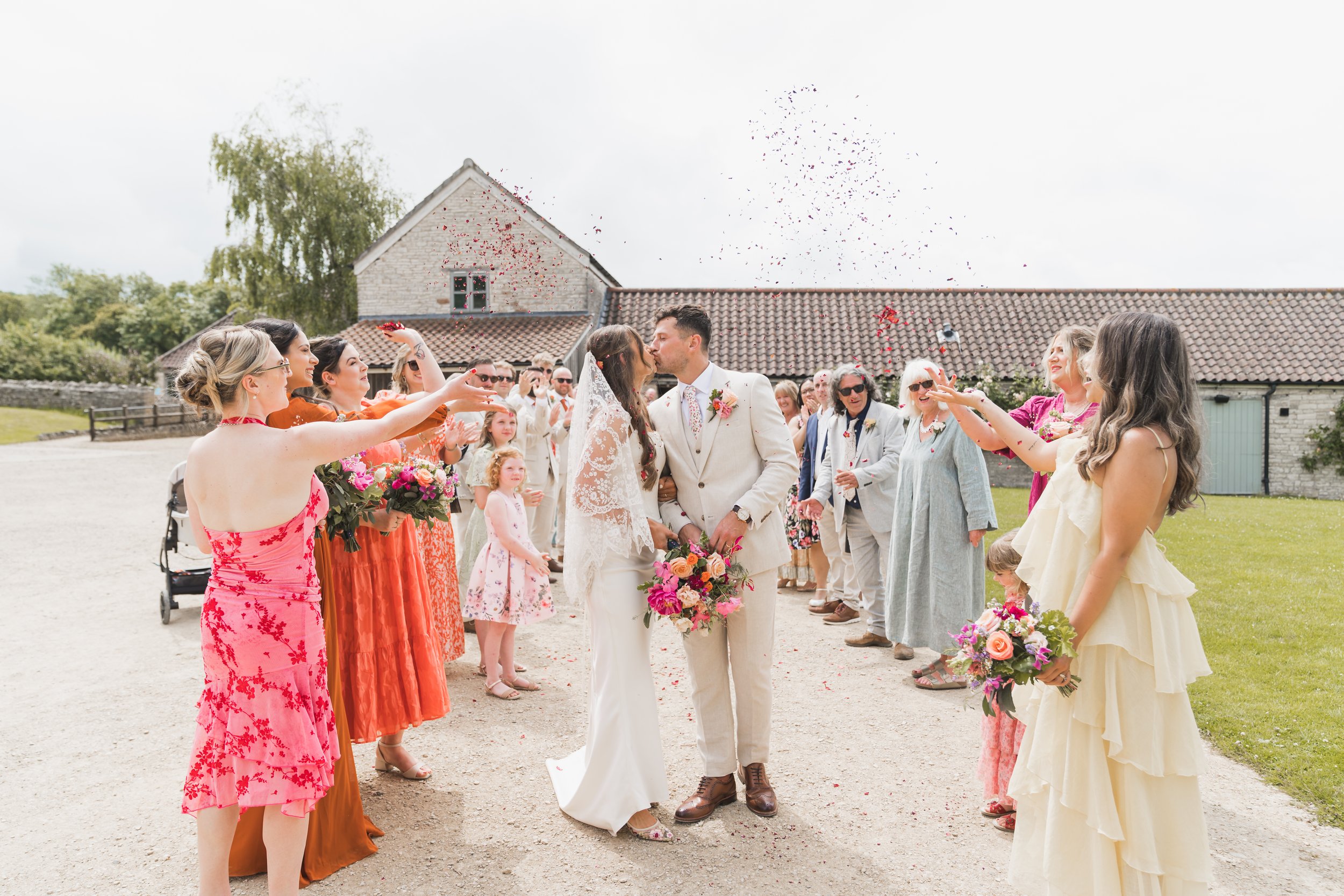 A group of women and a couple kiss during a wedding celebration outdoors, with friends and family celebrating with flower bouquets and confetti in the air.