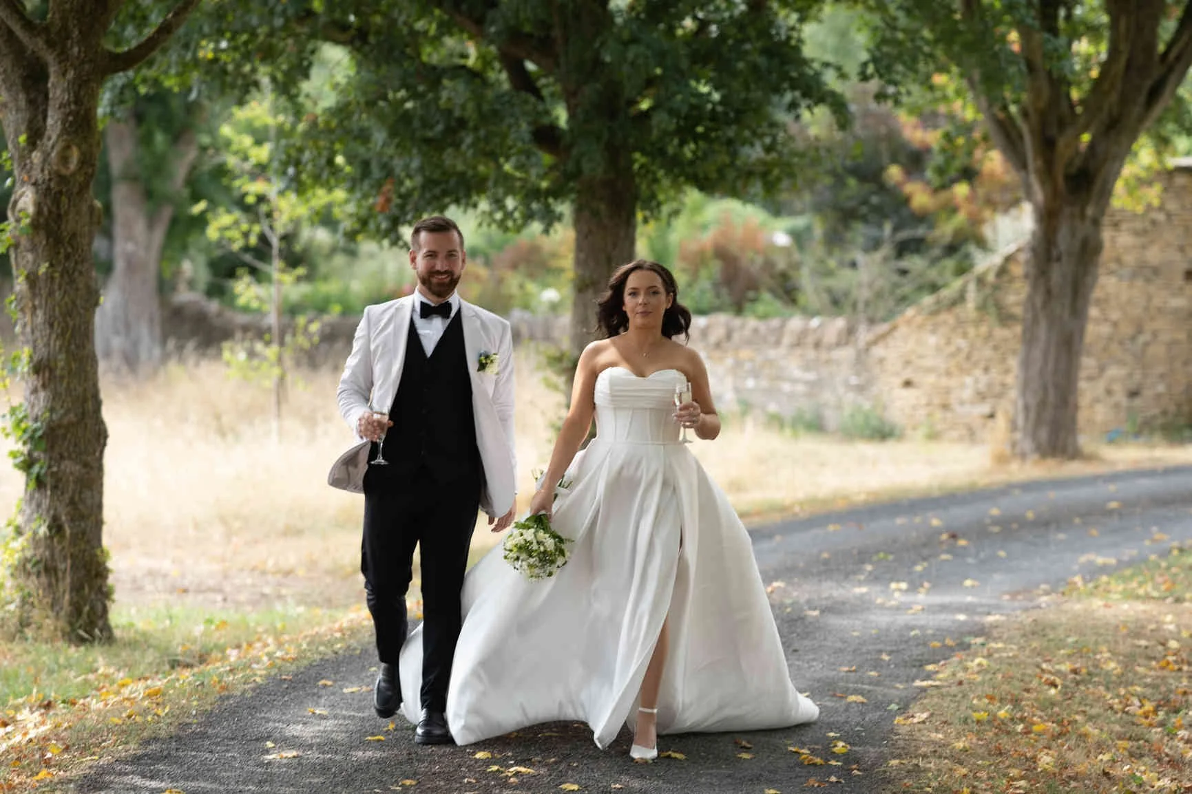 A bride and groom walking outdoors under trees on their wedding day. The bride is wearing a strapless white wedding gown and holding a bouquet and glass of champagne in Gloucester