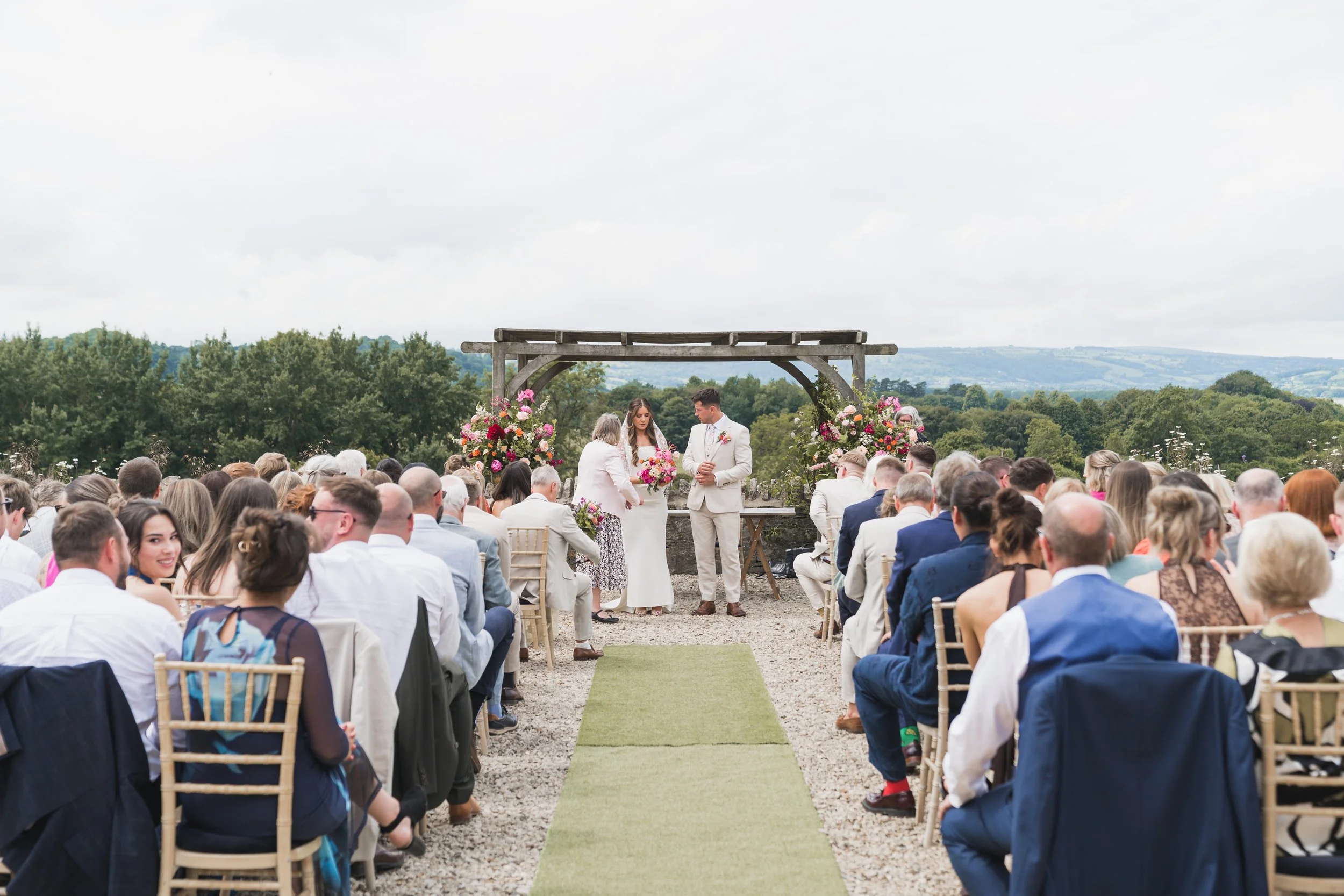 An outdoor wedding ceremony with a bride and groom standing under a wooden arch with floral arrangements, surrounded by seated guests, scenic countryside background with green trees and hills, overcast sky.