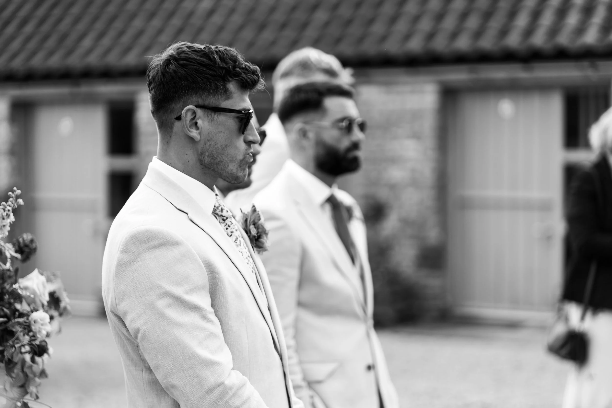 Black and white photo of two men standing in a line at an outdoor wedding ceremony, wearing sunglasses and suits, with a building in the background.