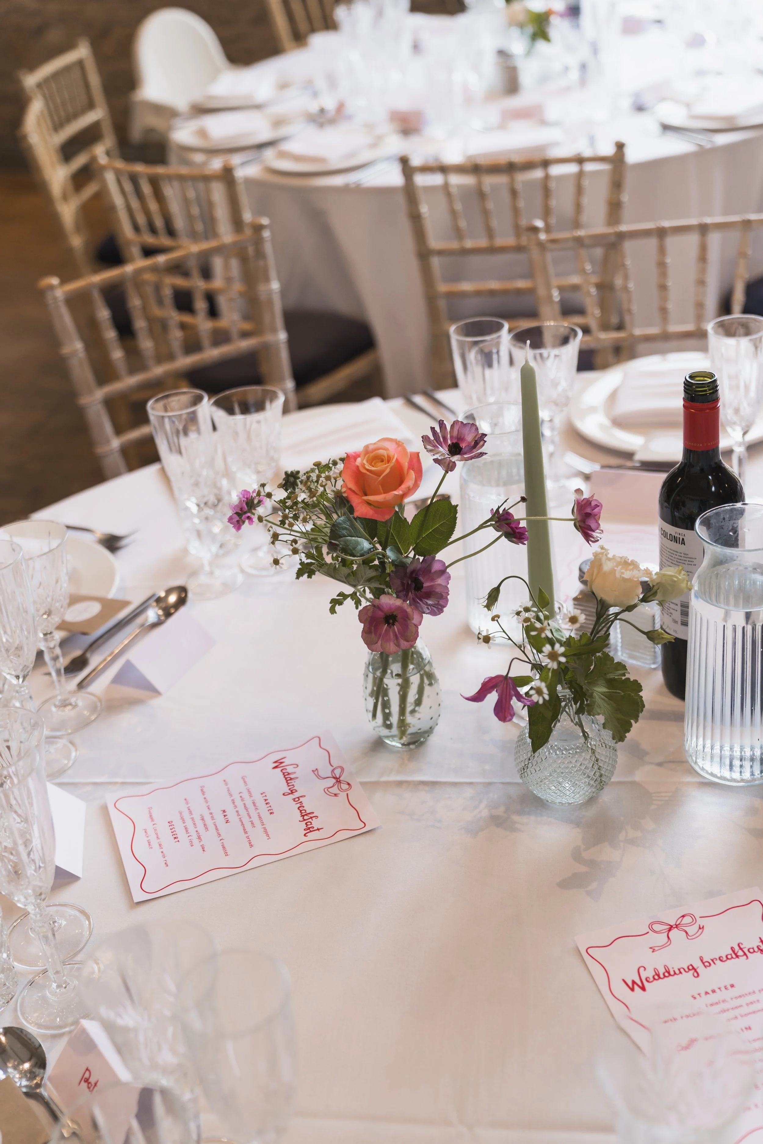 A wedding reception table with a floral centerpiece, wine, glasses, silverware, and a wedding breakfast menu, with elegant chairs surrounding the table.