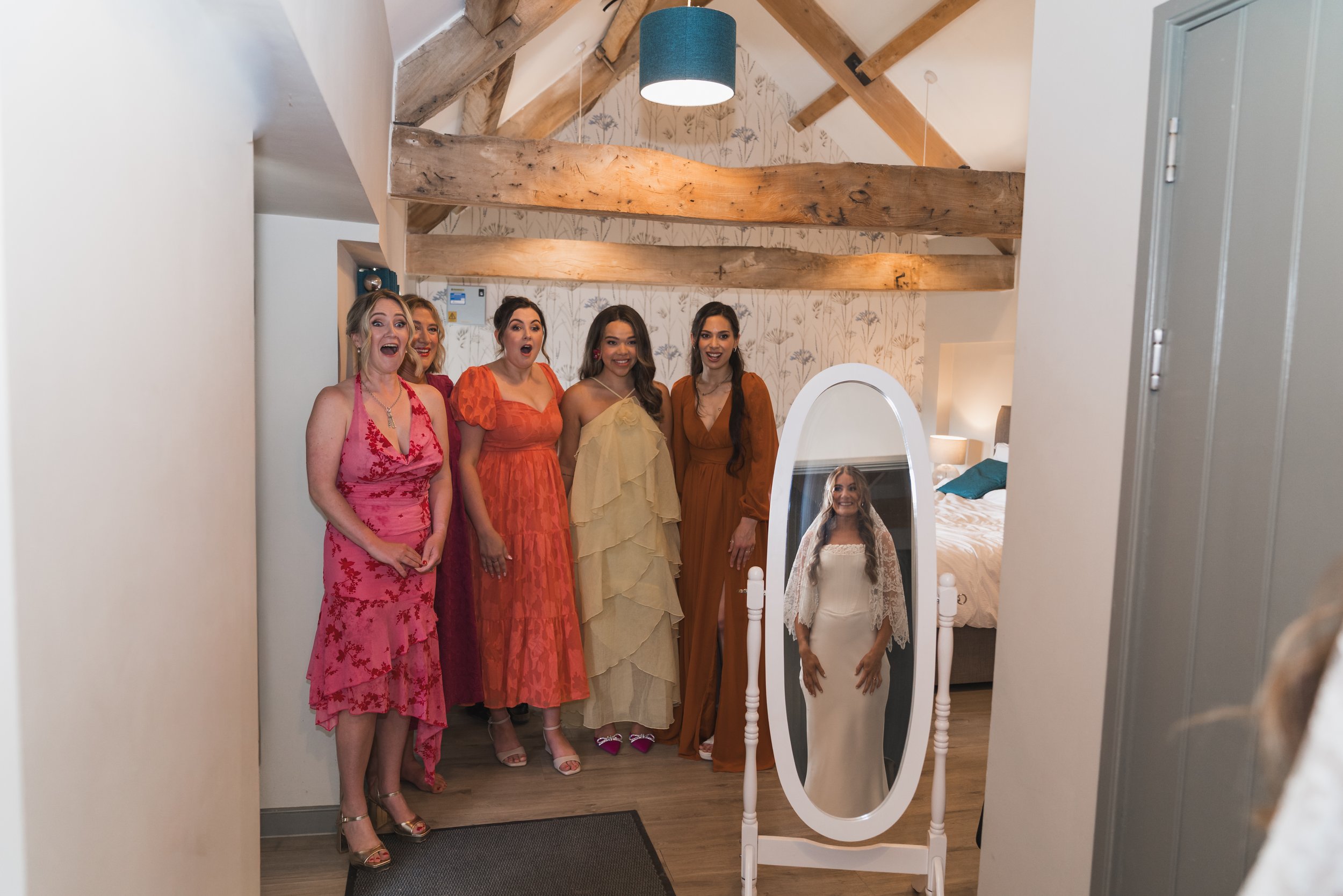 A group of five women dressed in colorful dresses standing in front of a mirror in a room with wooden beams and floral wallpaper. One woman is reflected in the mirror, wearing a white lace dress.