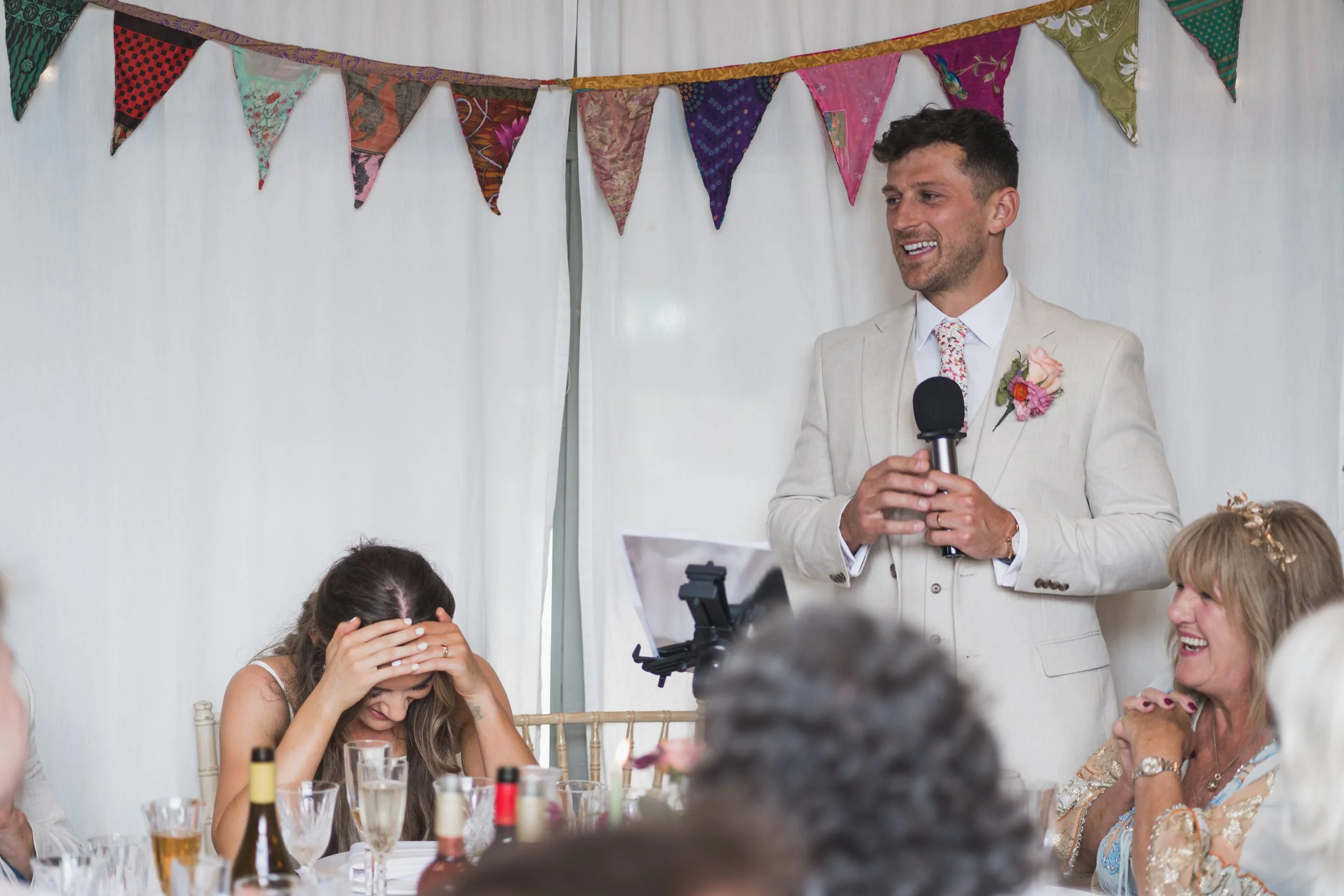A man in a light-colored suit holding a microphone, speaking at a celebration or wedding event. Several women are sitting at a table, some laughing and others covering their faces with their hands. Decorative colorful fabric bunting hangs behind, and a dining table is set with drinks and wine bottles.
