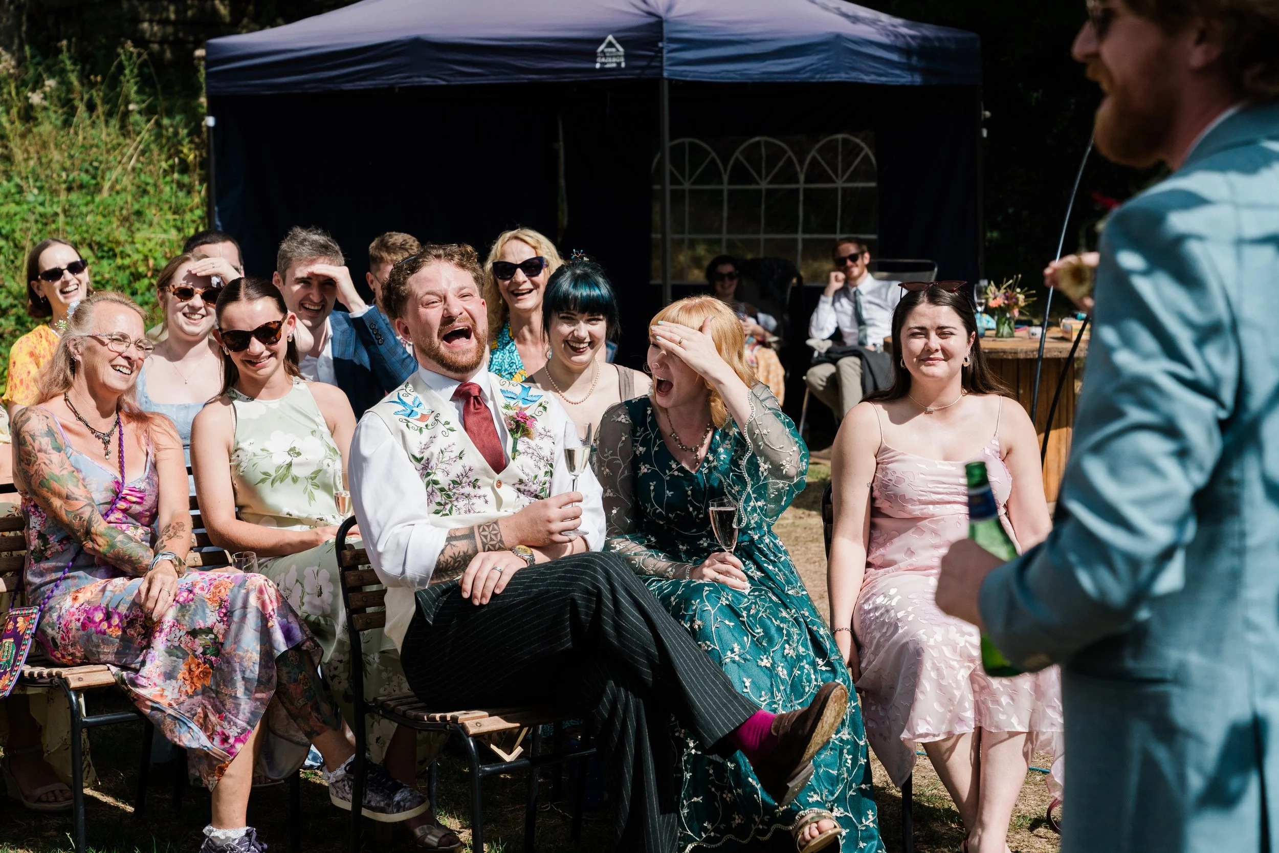 A group of people at an outdoor gathering or celebration, sitting and laughing, with a man in a vest and tie holding a wine glass in the center, and a person speaking or performing in front of them captured by Lapwing Farm wedding photographer