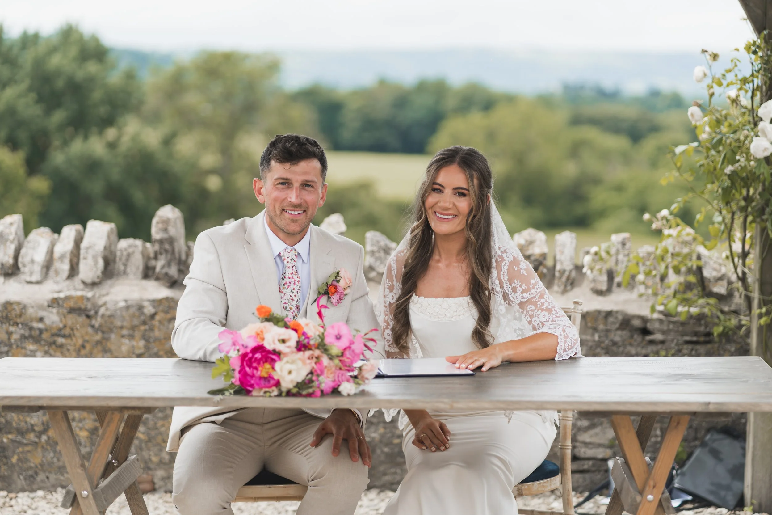 A wedding couple seated at a table outdoors, with a bouquet of pink and white flowers in front of them. The groom is dressed in a light-colored suit and tie, smiling at the camera. The bride is wearing a white lace dress with a veil, also smiling. The background features trees and a stone fence, indicating a scenic outdoor setting.