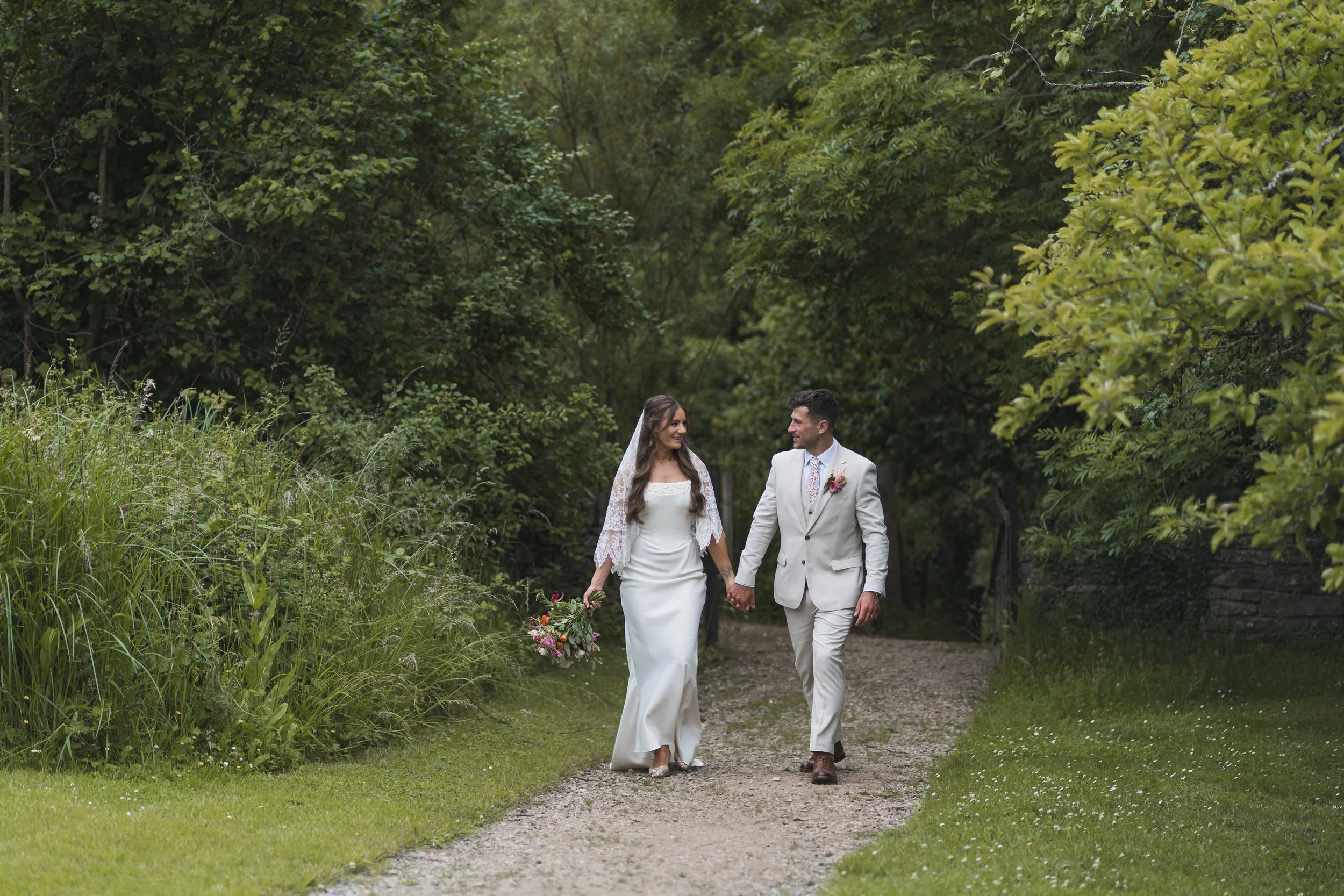 A bride and groom walking hand-in-hand on a forest trail, dressed in wedding attire, surrounded by green trees and plants.