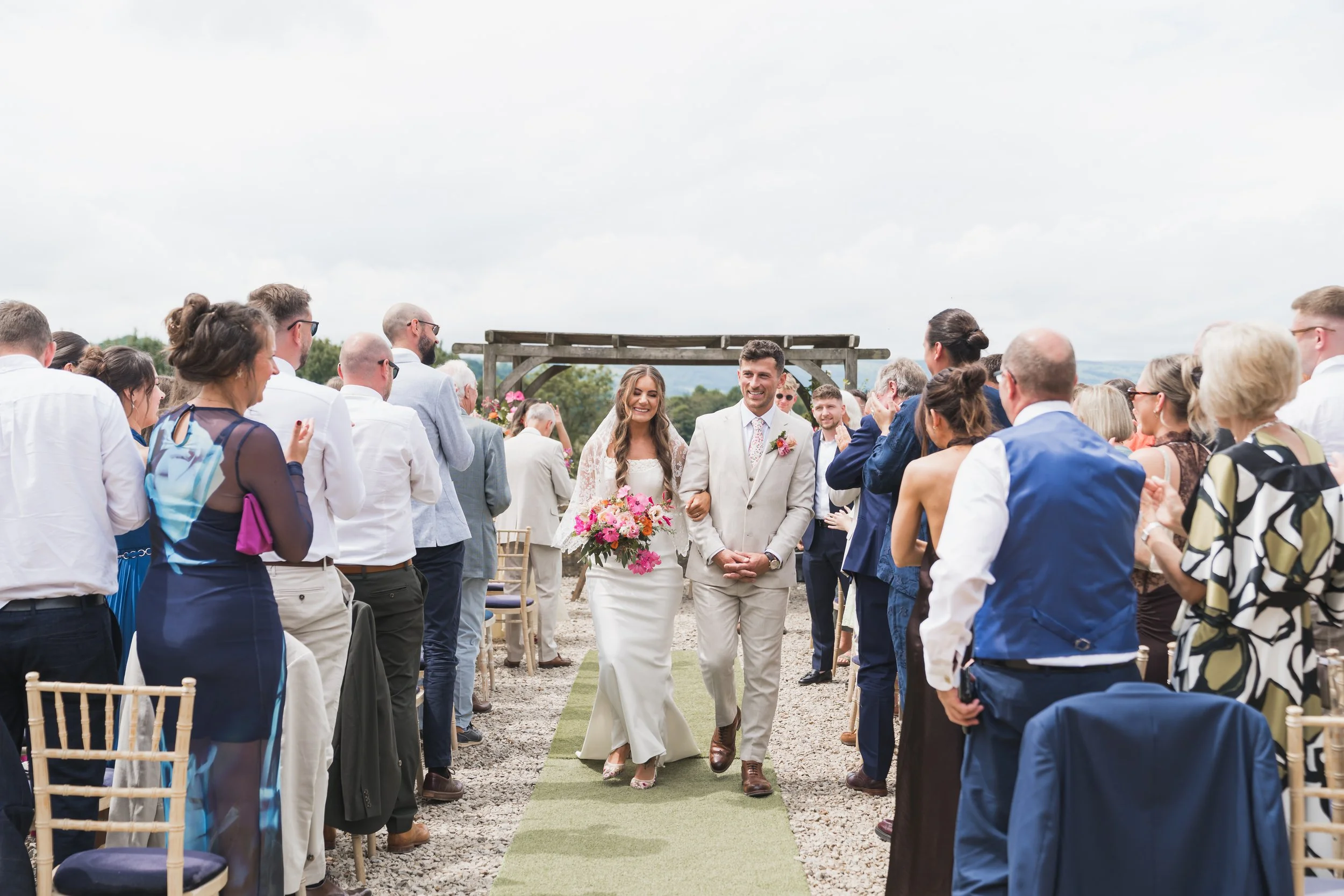 A wedding ceremony outdoors with a bride and groom walking down the aisle, surrounded by seated guests on either side. The bride is holding a bouquet and smiling, while the groom is in a suit, also smiling. The setting appears to be rustic with an open sky and countryside in the background.