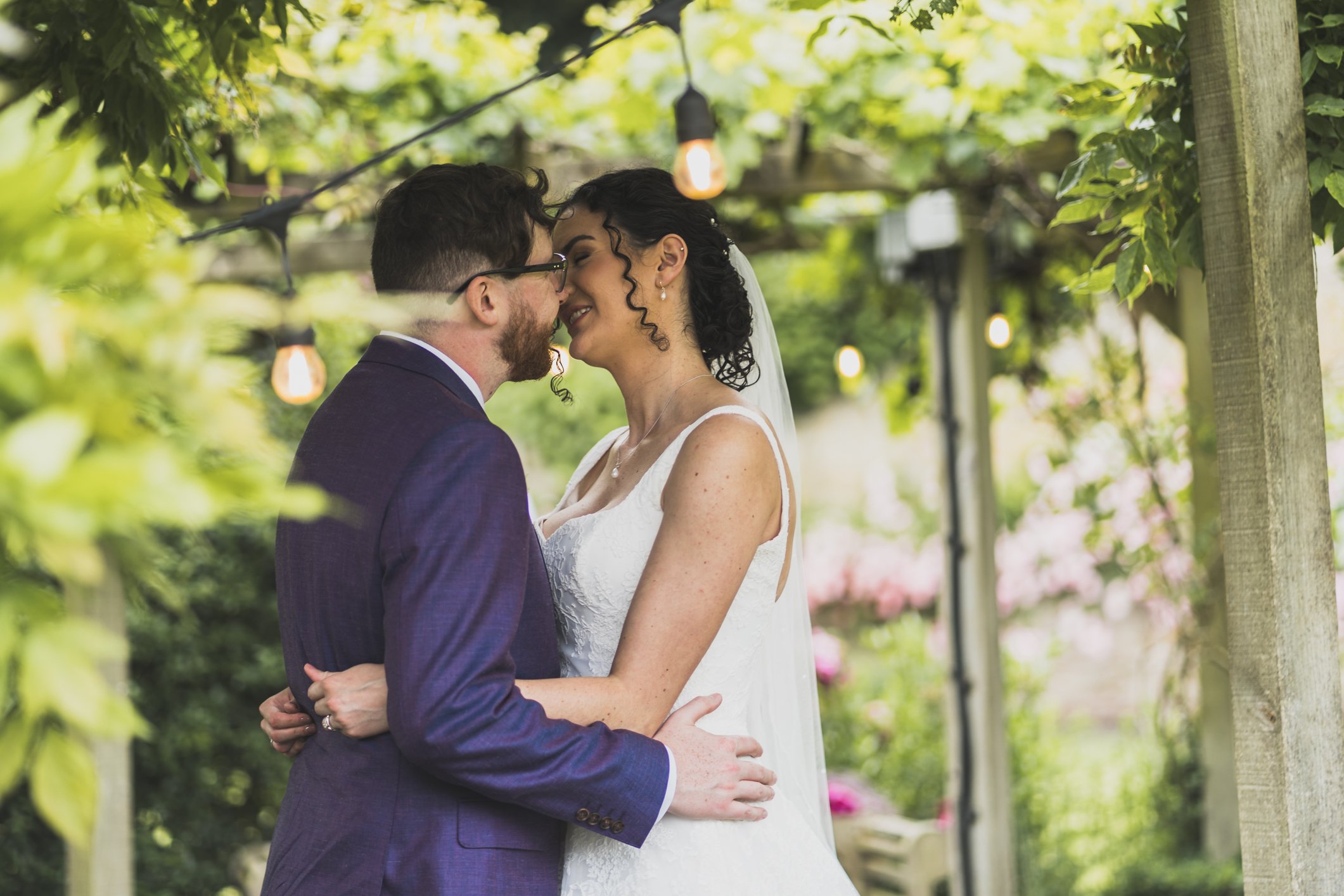 A bride and groom sharing a kiss under a tree with hanging string lights during their outdoor weddingat Priston Mill
