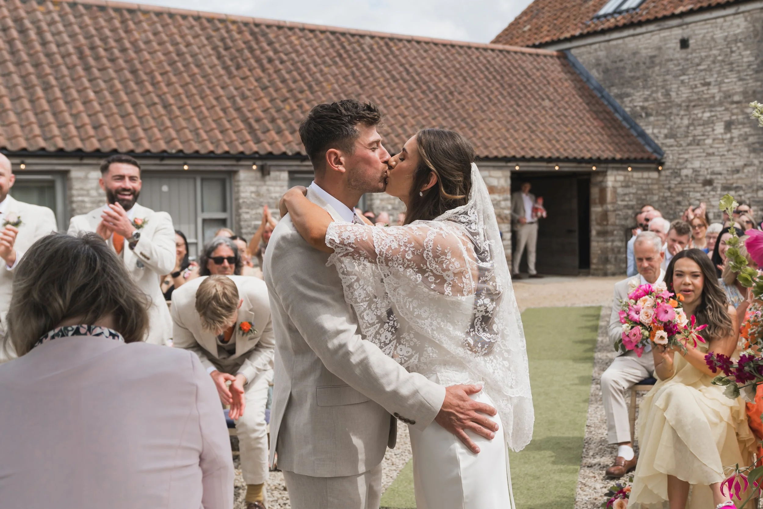 A bride and groom kiss during their outdoor wedding ceremony with guests seated and clapping, in front of a rustic brick building with a tiled roof and string lights.