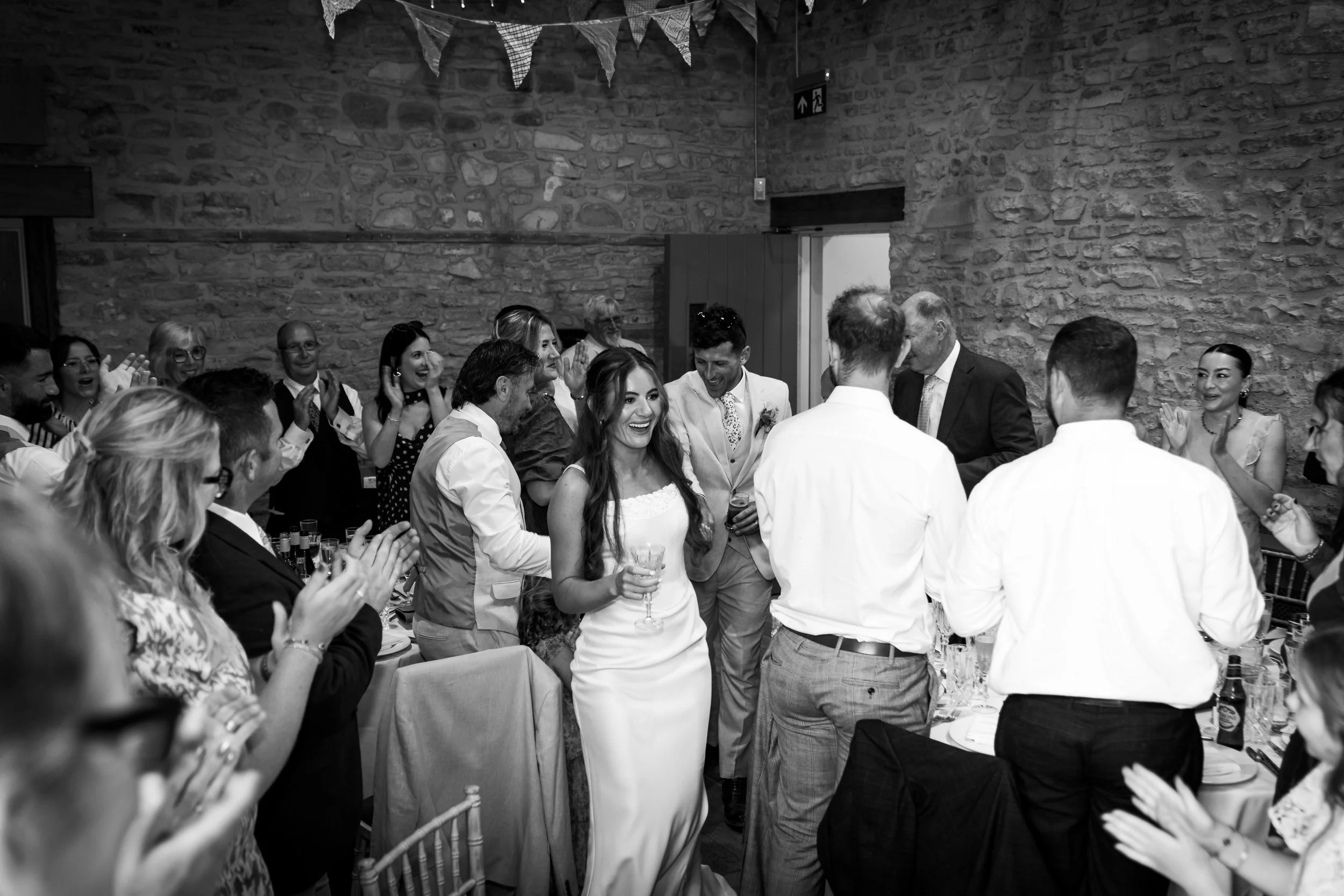 A black and white photo of a group of people gathered at a celebration or party, with some clapping, smiling, and holding drinks, against a rustic stone wall with bunting decorations.