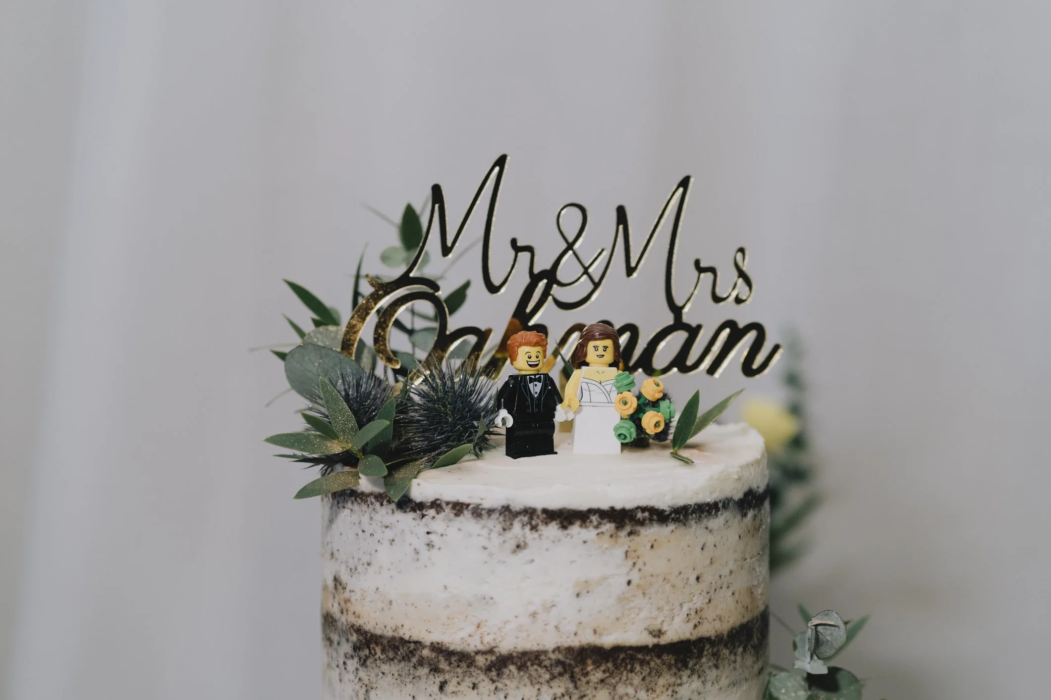Wedding cake decorated with green leaves, black flowers, and LEGO bride and groom figurines, topped with a 'Mr & Mrs' cake topper.