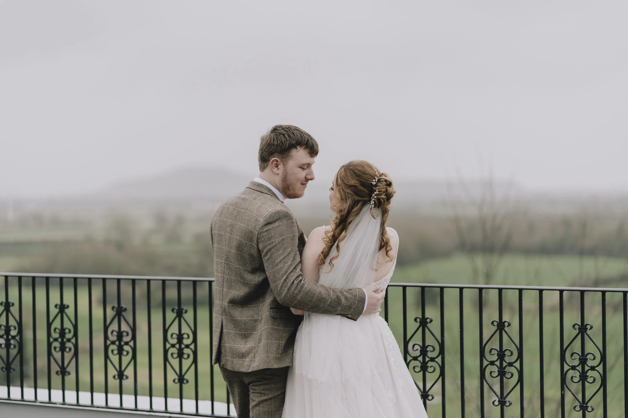 A newlywed couple stands close, gazing into each other's eyes, on a balcony overlooking Mendips with a scenic, blurred landscape background.