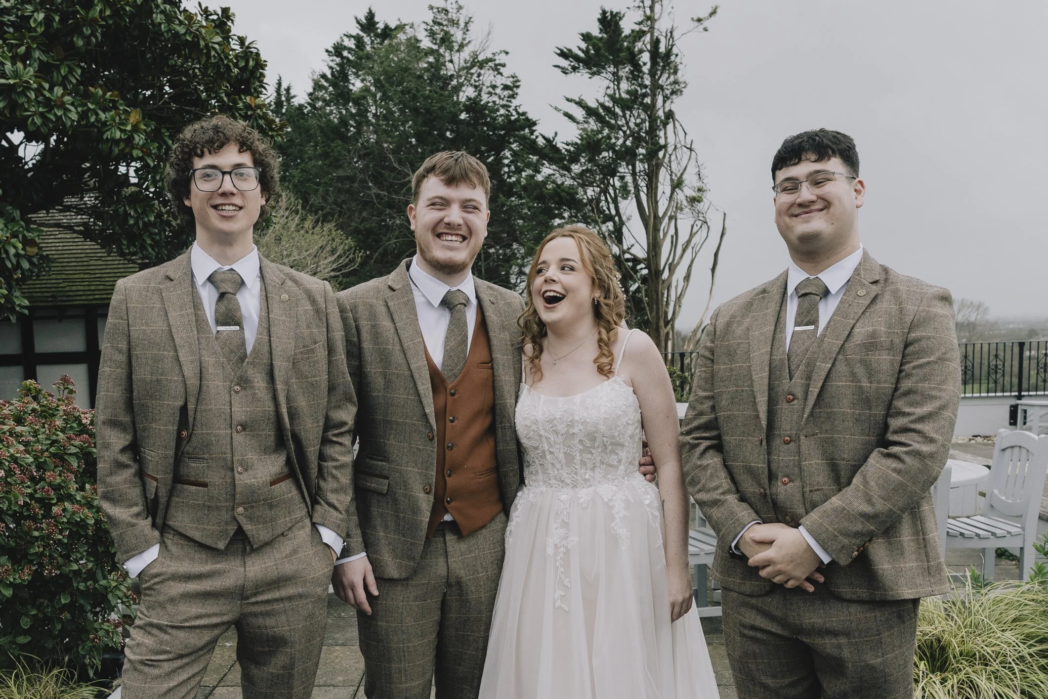 Groomsmen and Bride, dressed in vintage-style brown checkered suits and a white wedding gown, standing outdoors on a cloudy day, smiling and laughing, with trees and patio furniture in the background.
