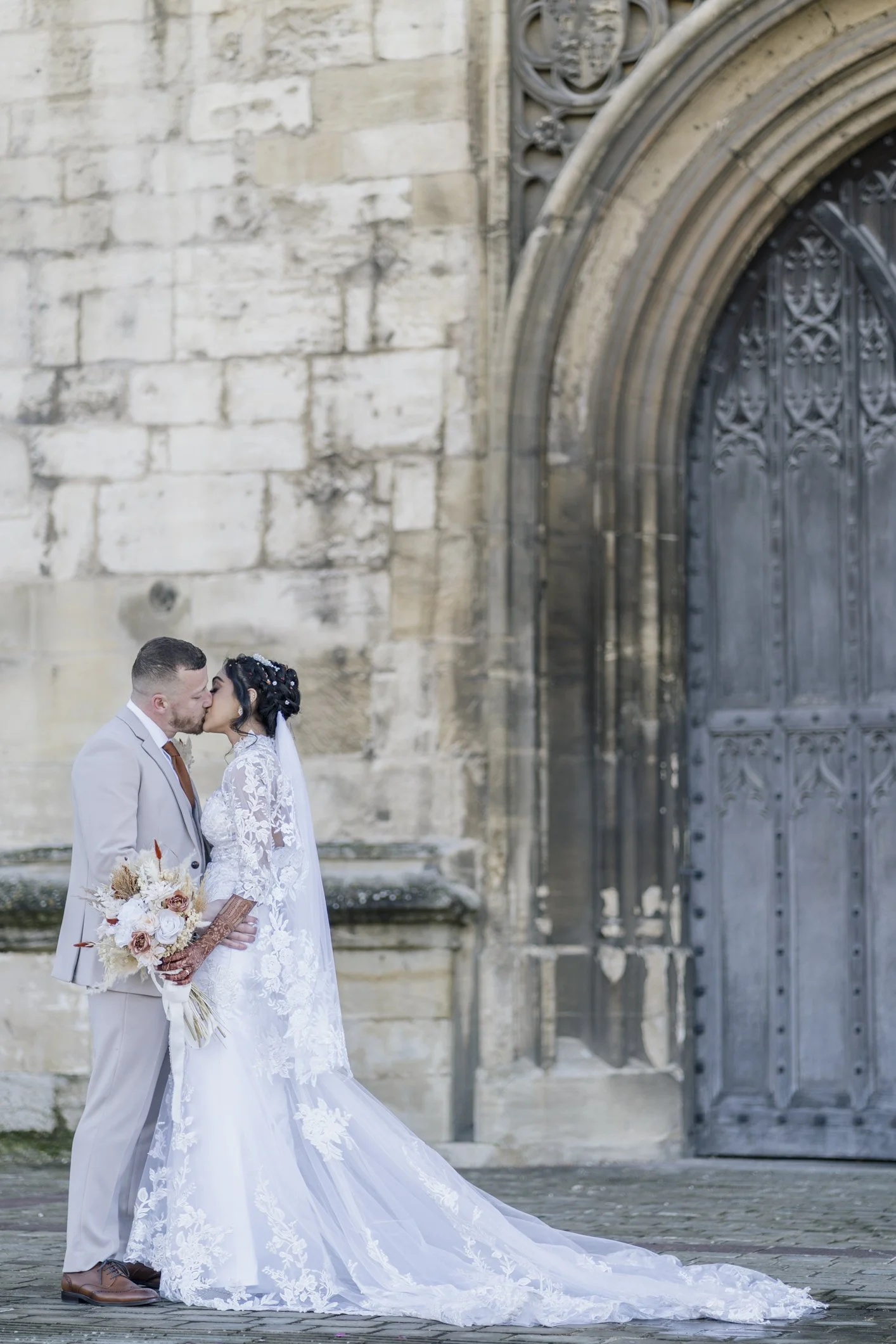 Wedding couple kissing in front of a historic stone building and large wooden door, with the bride wearing a lace wedding gown and veil, and holding a floral bouquet in Gloucestershire