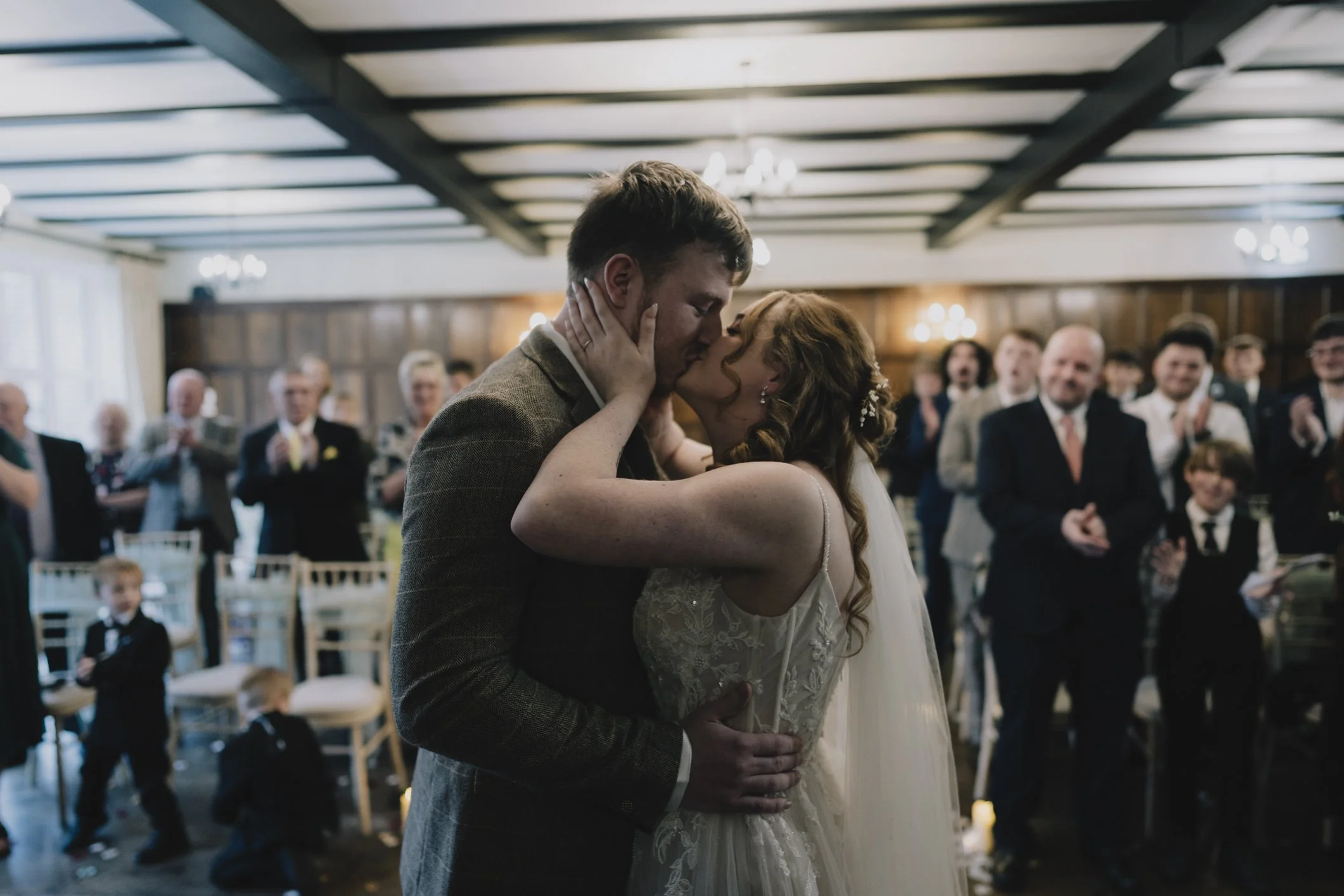 A bride and groom share a kiss during their wedding ceremony, surrounded by guests in the ceremony room at Webbington