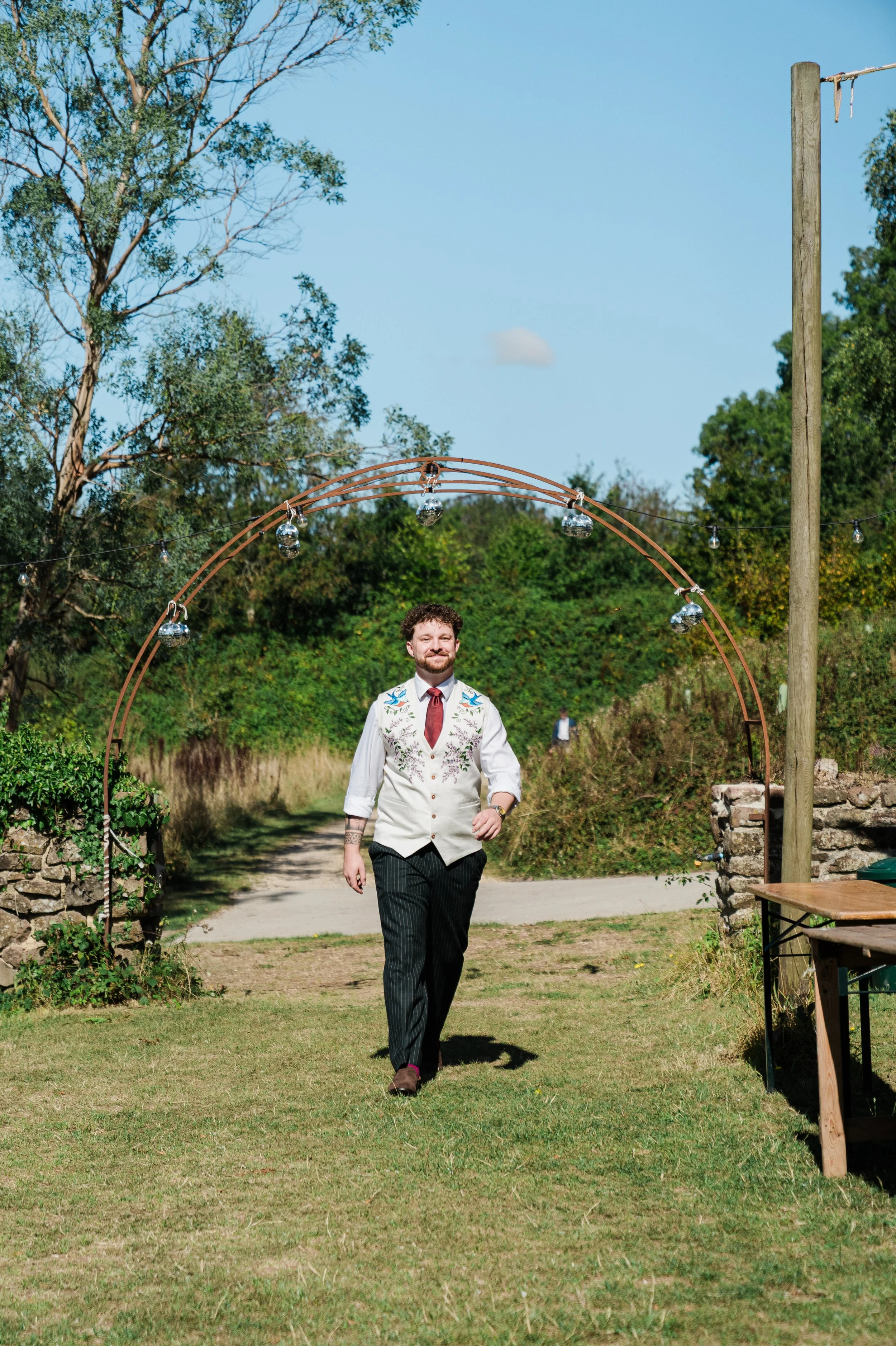The groom with a beard and curly hair walking on a grassy area outdoors, dressed in a white shirt with floral embroidery, striped pants, and brown shoes, with a decorative arch and trees in the background.