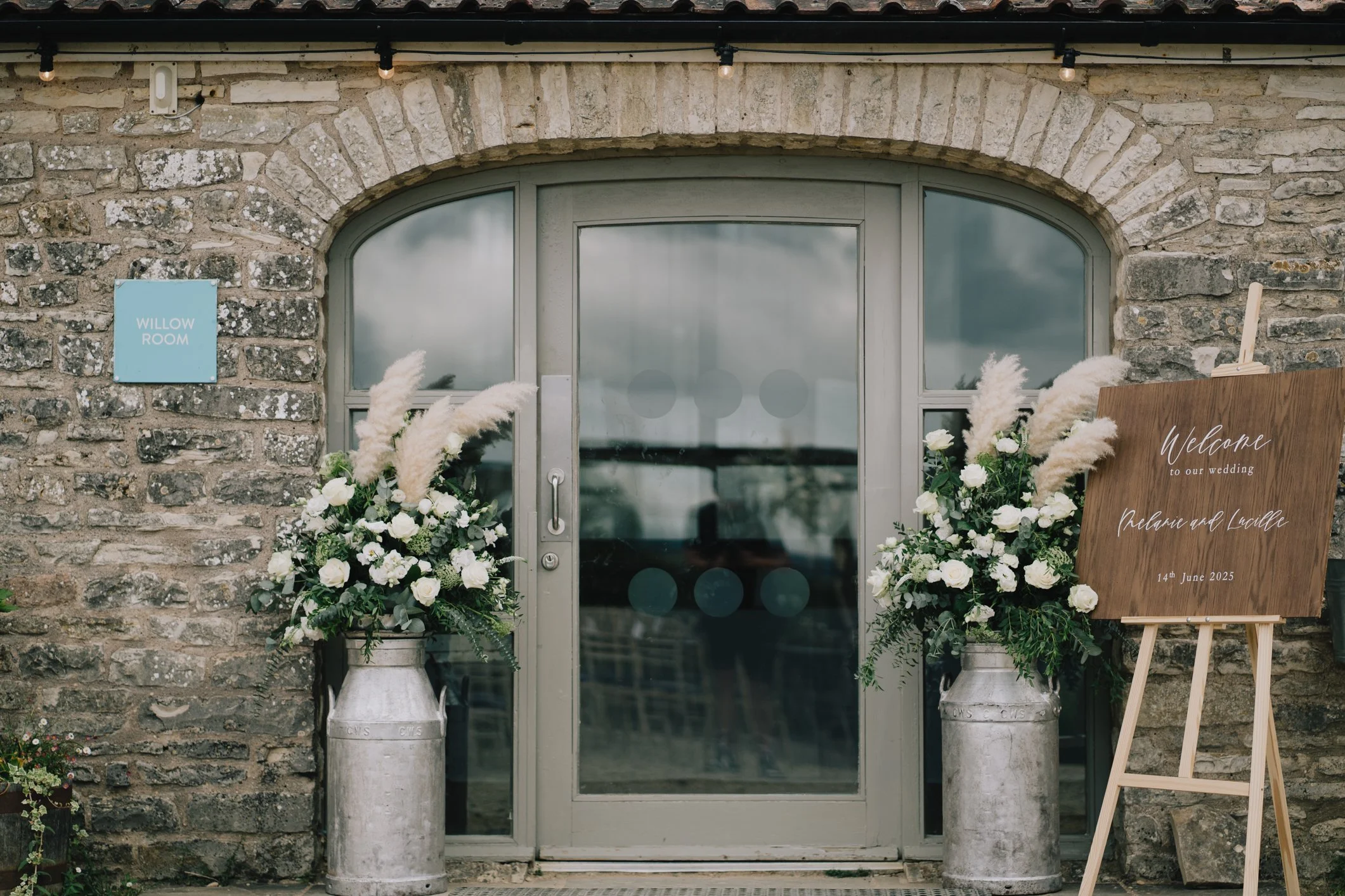 A stone building entrance with a gray double door, decorated for a wedding with two large floral arrangements of white roses and pampas grass on either side. A wooden sign reads 'Welcome to our wedding Melanie and Nicole 14th June 2025' and a blue sign says 'WILLOW ROOM'. String lights hang above the entrance.