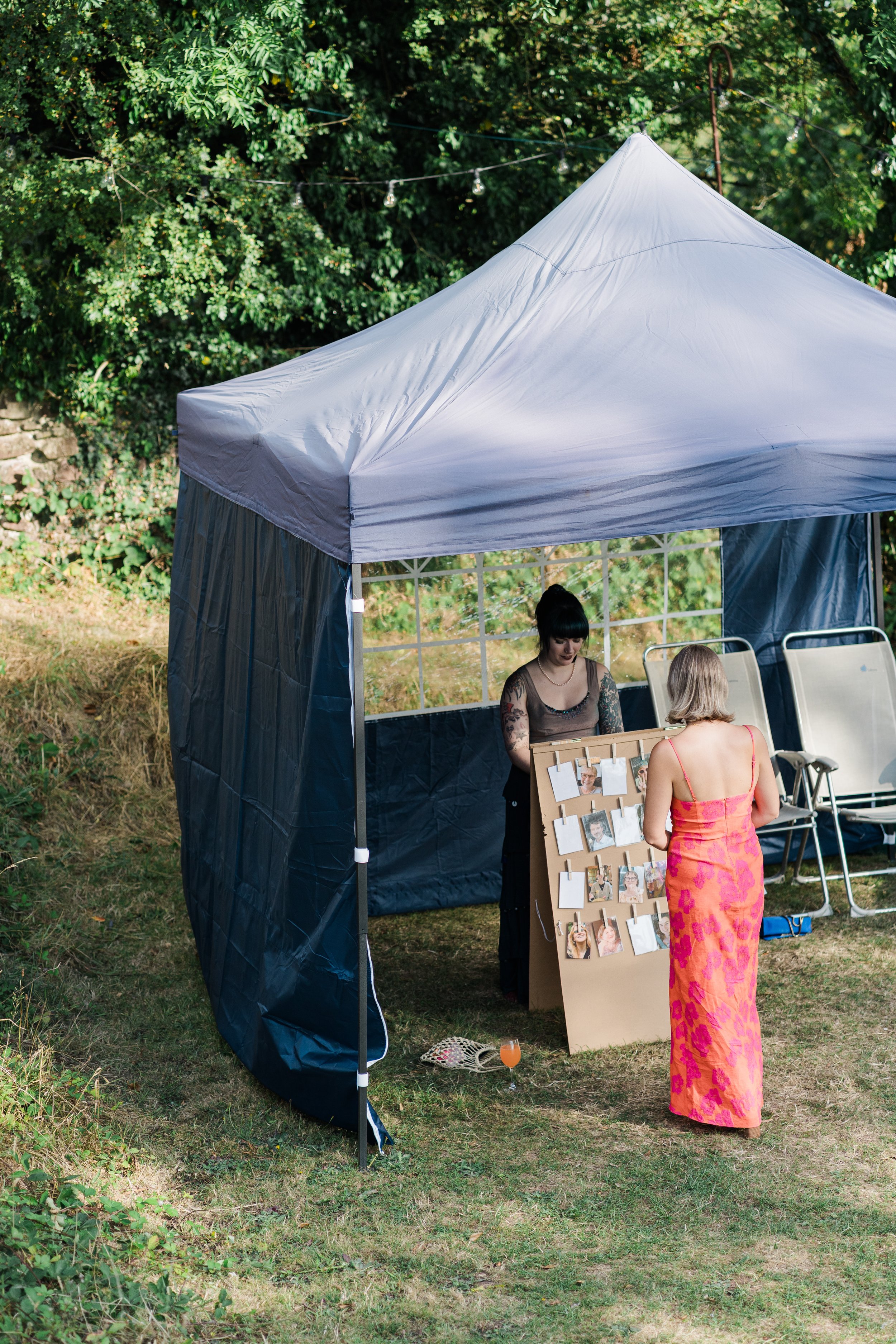 A woman in a pink and orange dress stands in front of a photo display at an outdoor event, talking to another woman inside a blue canopy tent. The tent is set up on a grassy area with trees and string lights in the background.