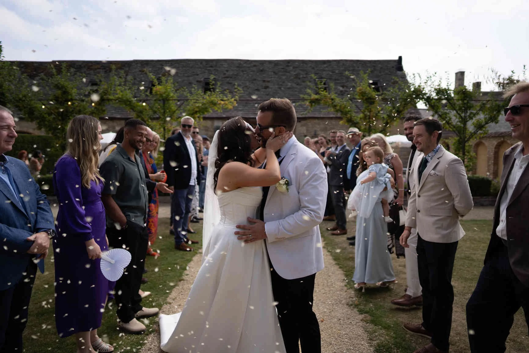 A bride and groom kiss during their outdoor wedding celebration as guests watch and celebrate around them, with white confetti falling in the air at Oxleaze Barn