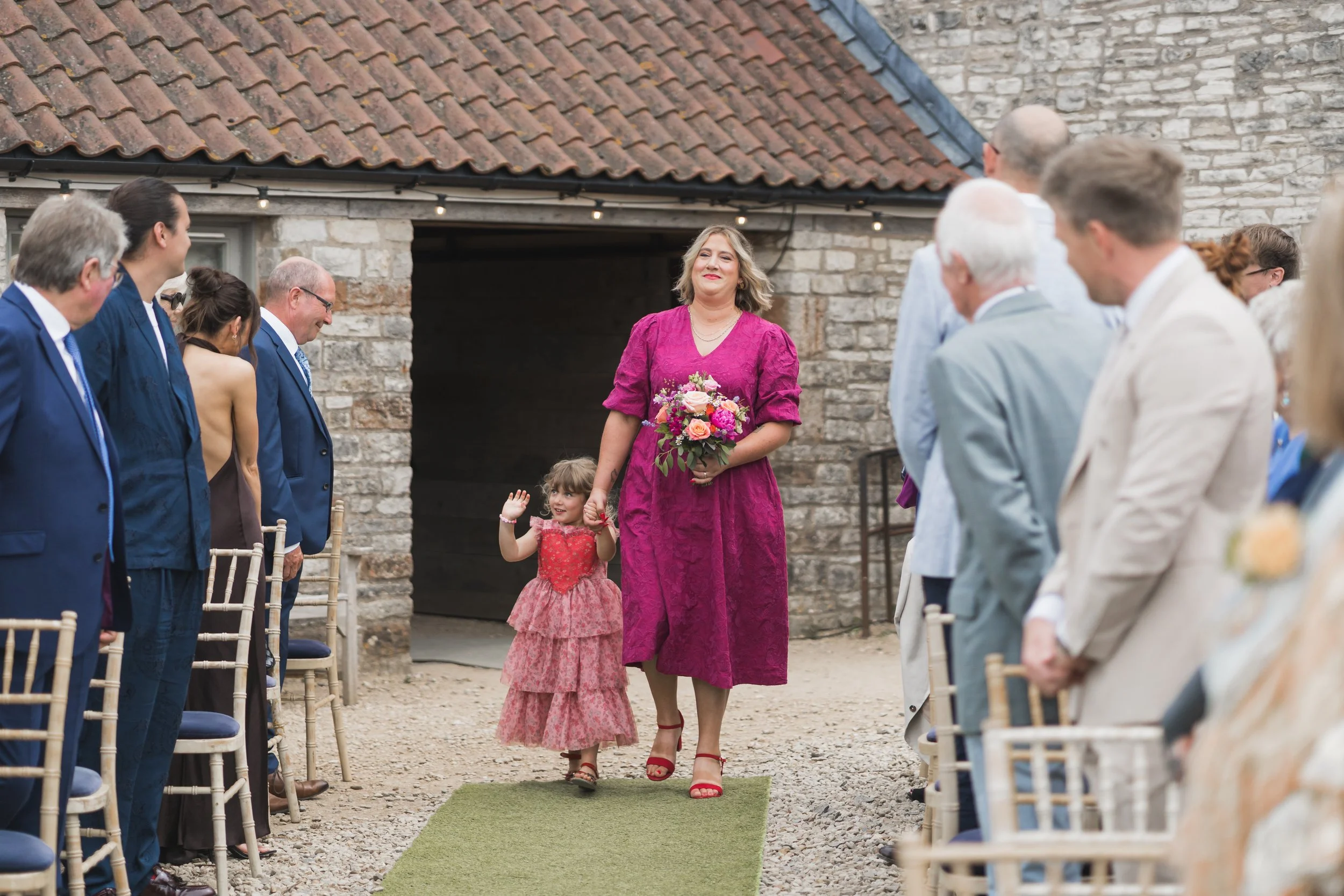 A woman in a pink dress holding a bouquet of flowers walks down the aisle hand-in-hand with a young girl in a pink dress, as guests seated on either side look on during a wedding ceremony outside in front of a stone building with a red-tiled roof.